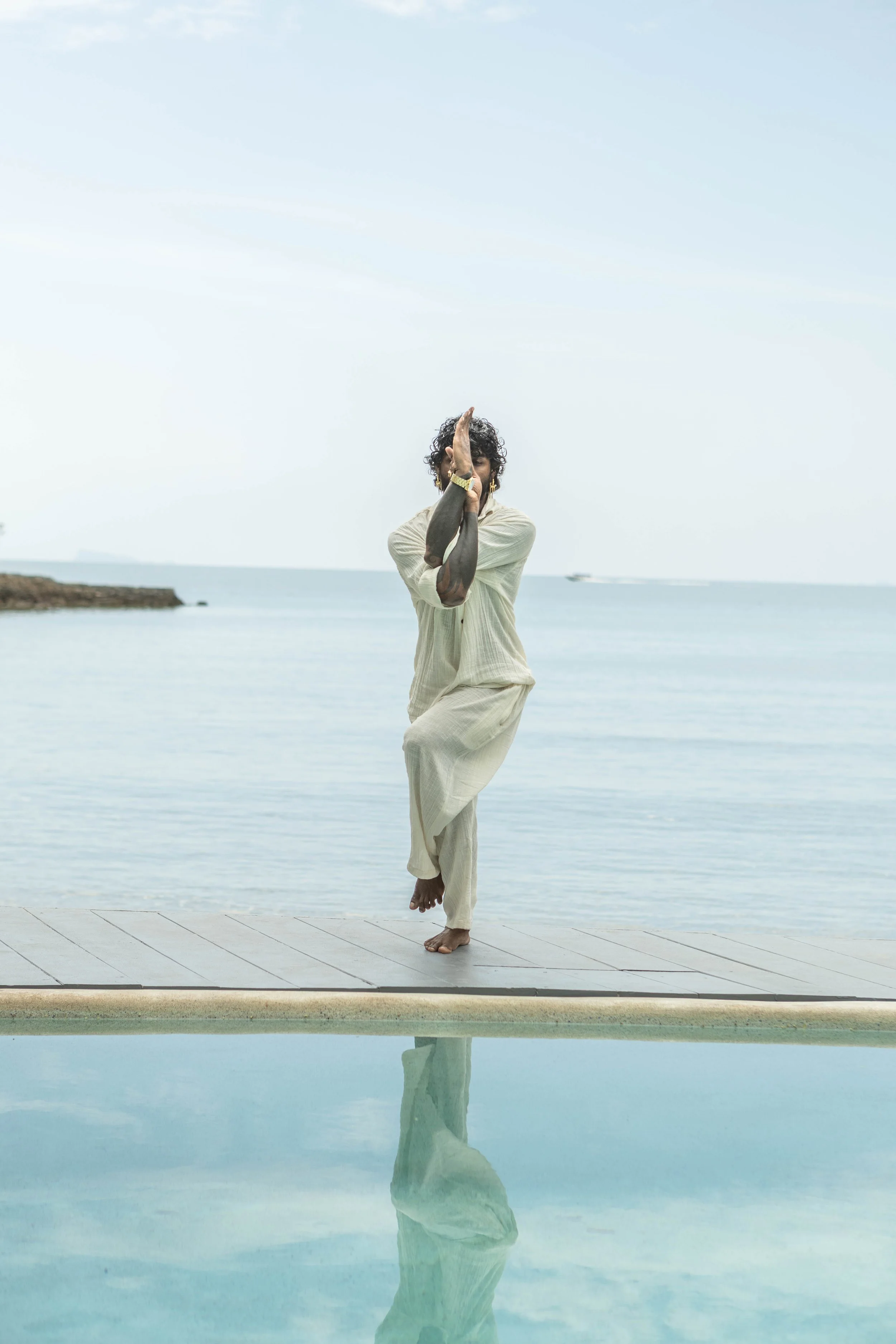 A person practicing yoga on a deck near the water with a calm ocean background, wearing light-colored clothing.