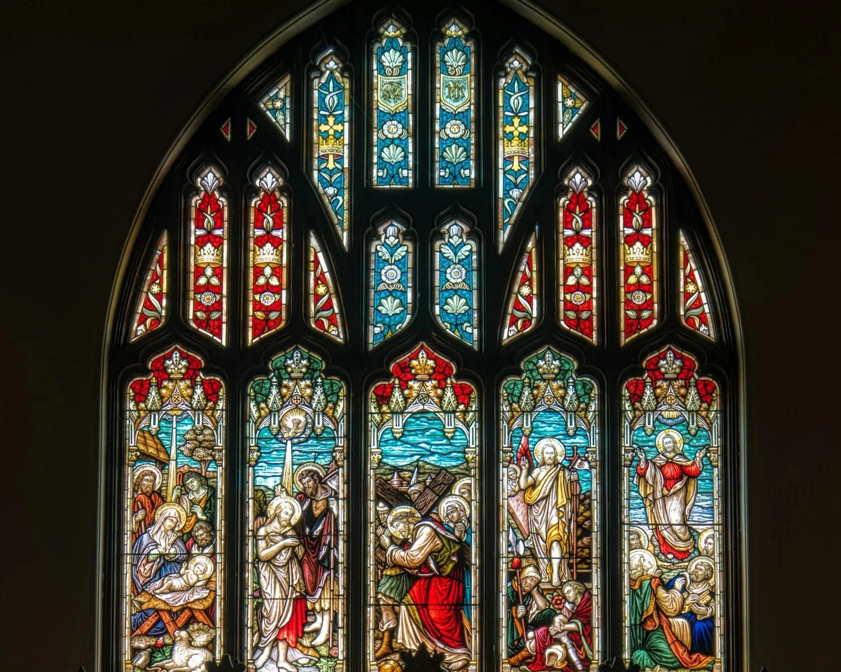 A large stained glass church window with religious scenes and figures, featuring intricate colorful designs and patterns.