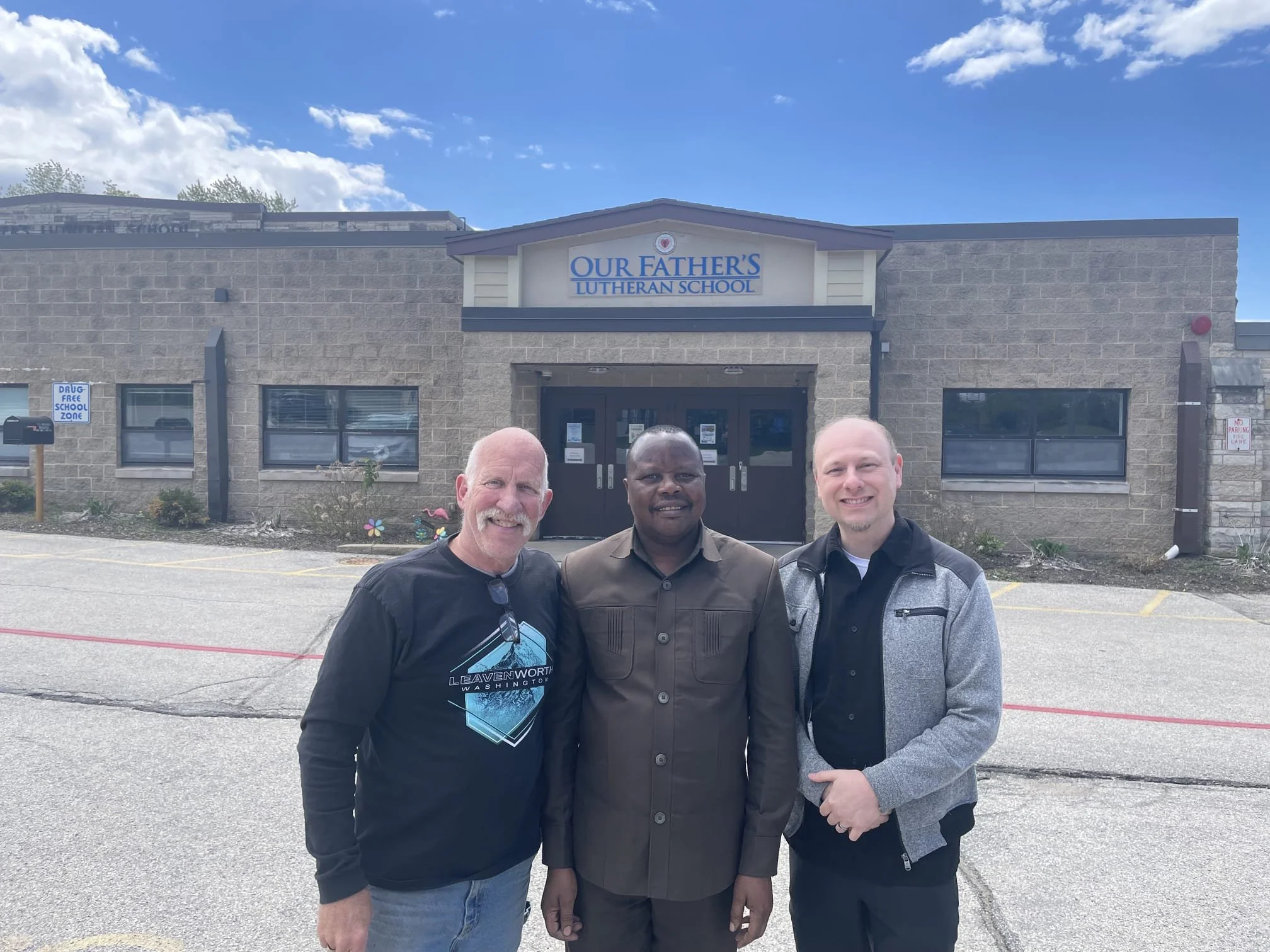 Three men standing outside a school building named Our Father's Lutheran School, with a blue sky and clouds above.