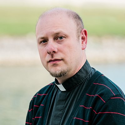 A man in clergy attire with a clerical collar, standing outdoors with a blurred natural background.