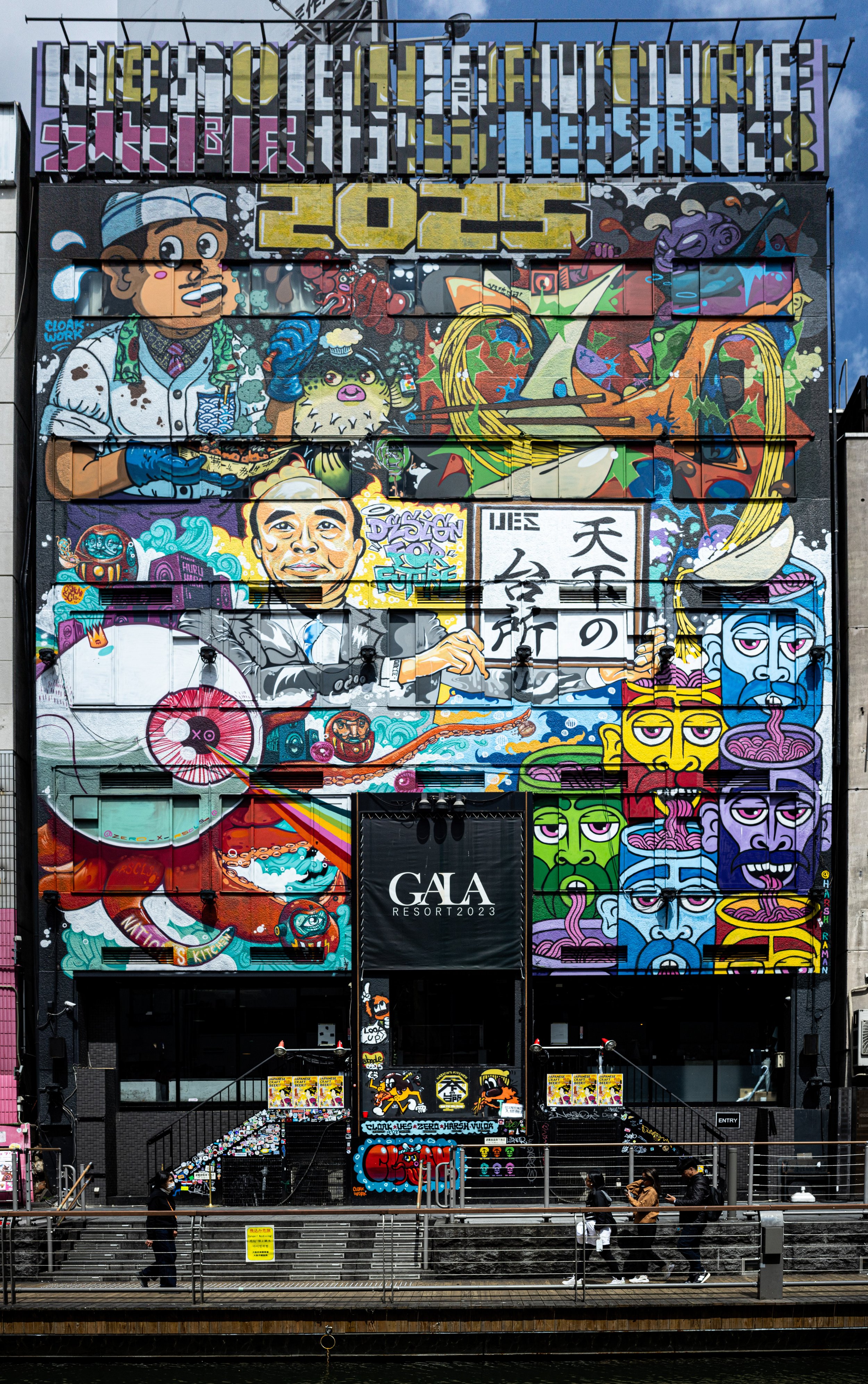 Colorful graffiti mural on a building featuring various characters, Japanese calligraphy, and abstract designs, with people walking in front.