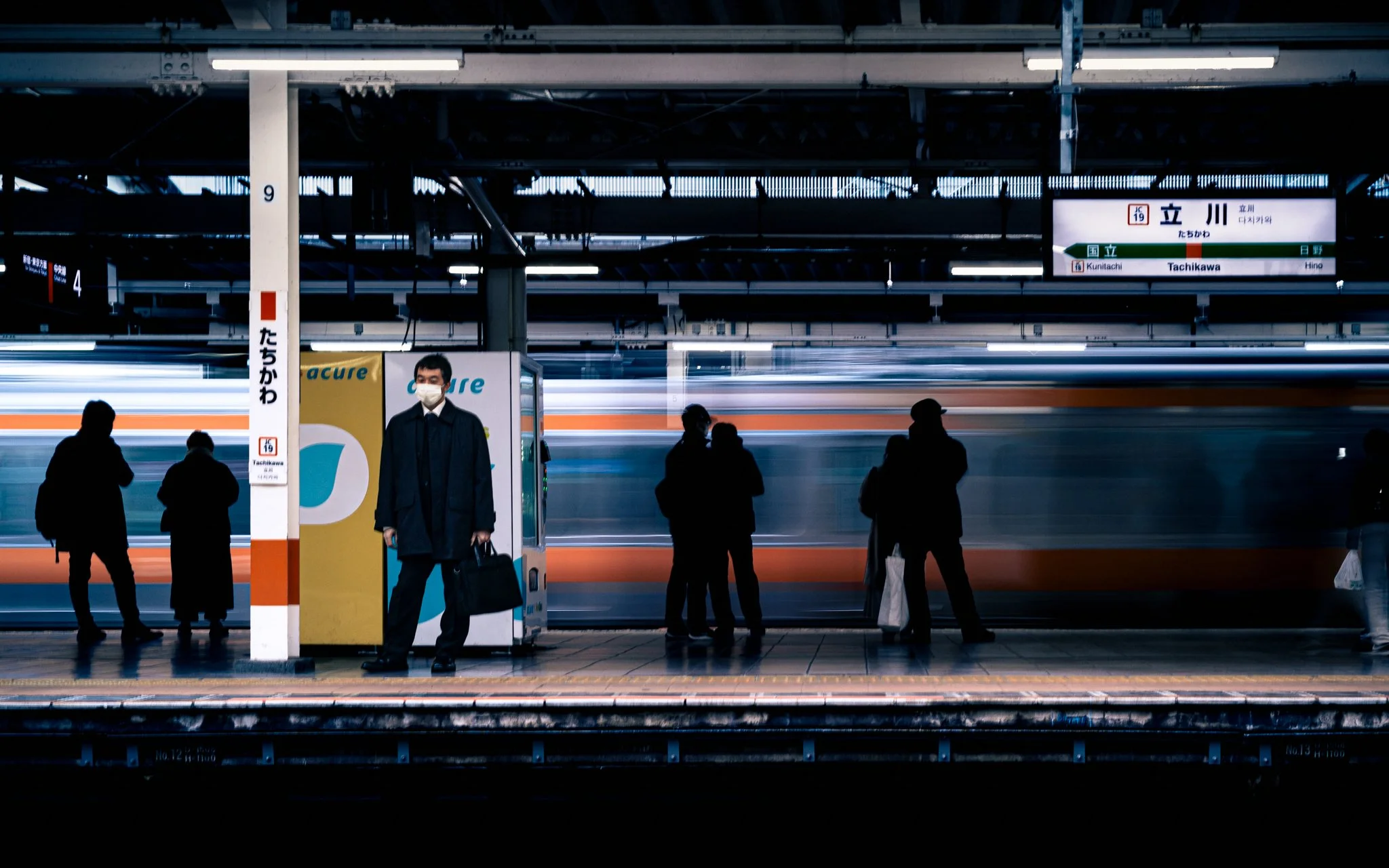 People waiting on a train platform at Takashima station, with an incoming train blurred in motion, and signs indicating station names in Japanese and English.