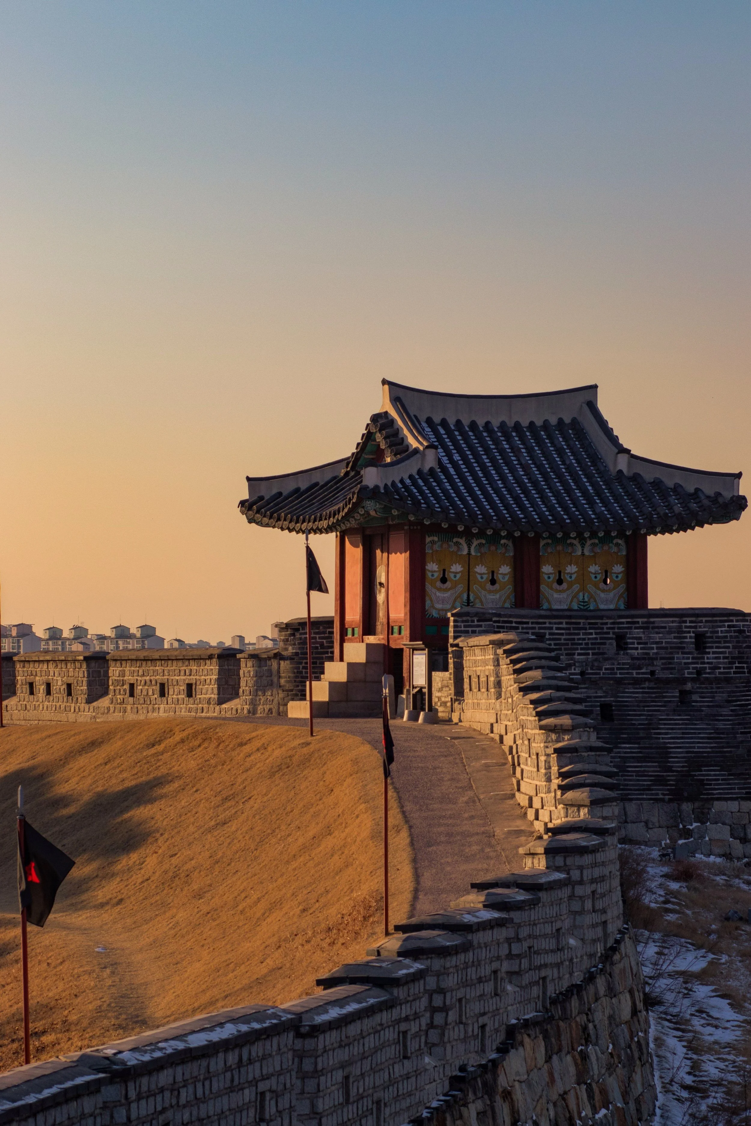 Traditional Korean fortress gate with colorful painted wooden details and a curved tiled roof, located atop a stone wall at sunset.
