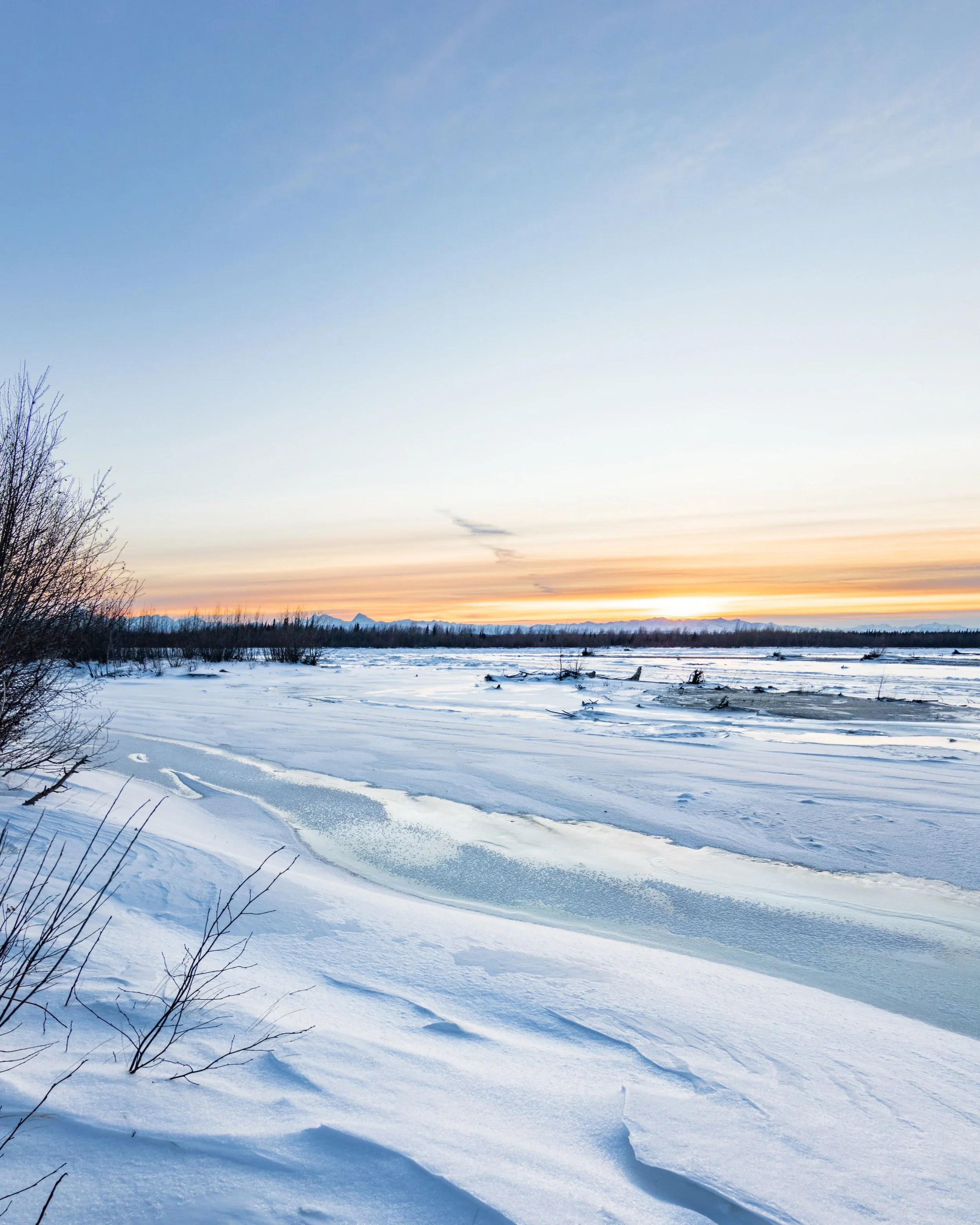 Snow-covered landscape with frozen river, bare trees on the left, and distant mountains at sunset.