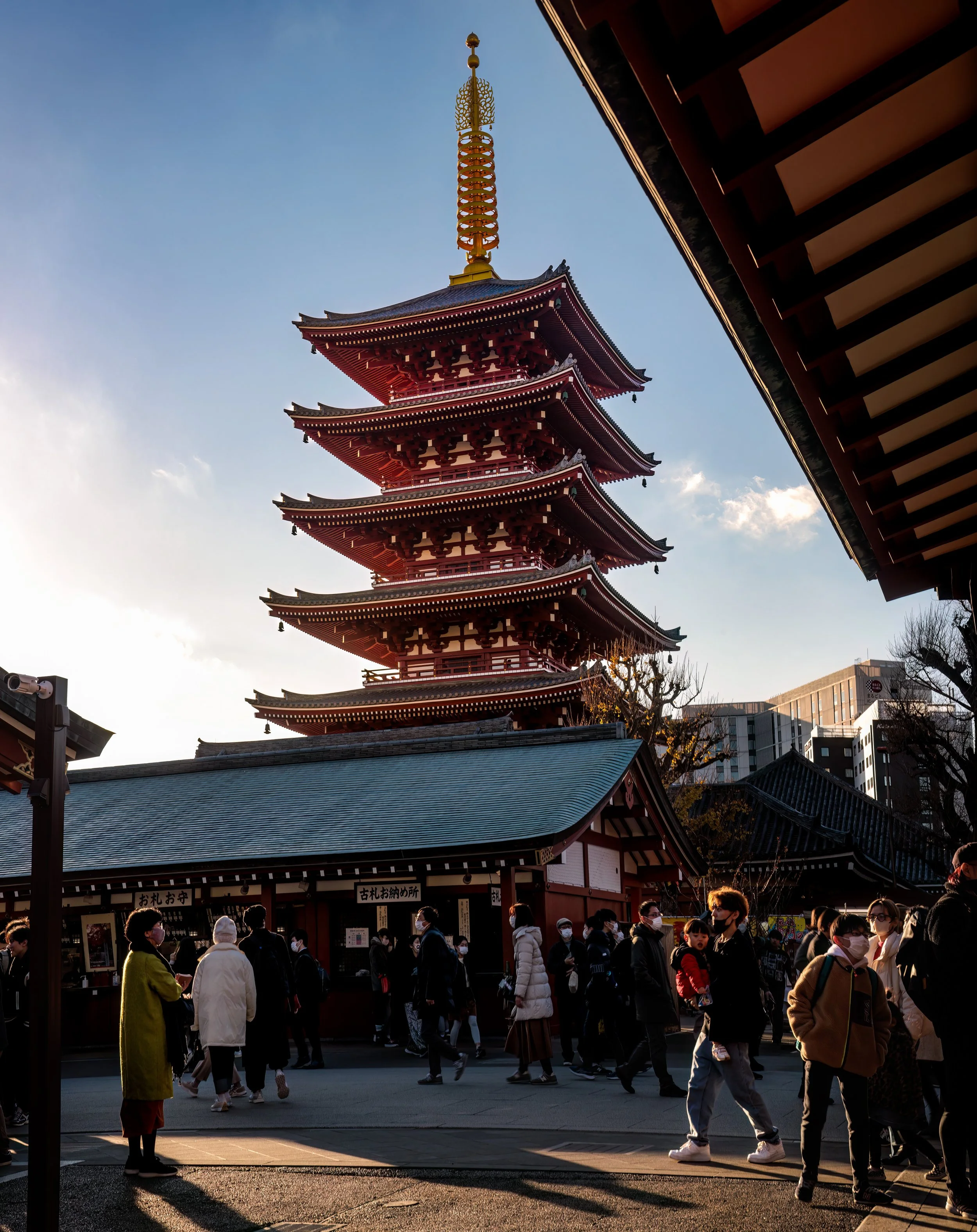 Crowded street scene outside a traditional Japanese pagoda with multiple tiers, surrounded by modern buildings, during late afternoon with sunlight.