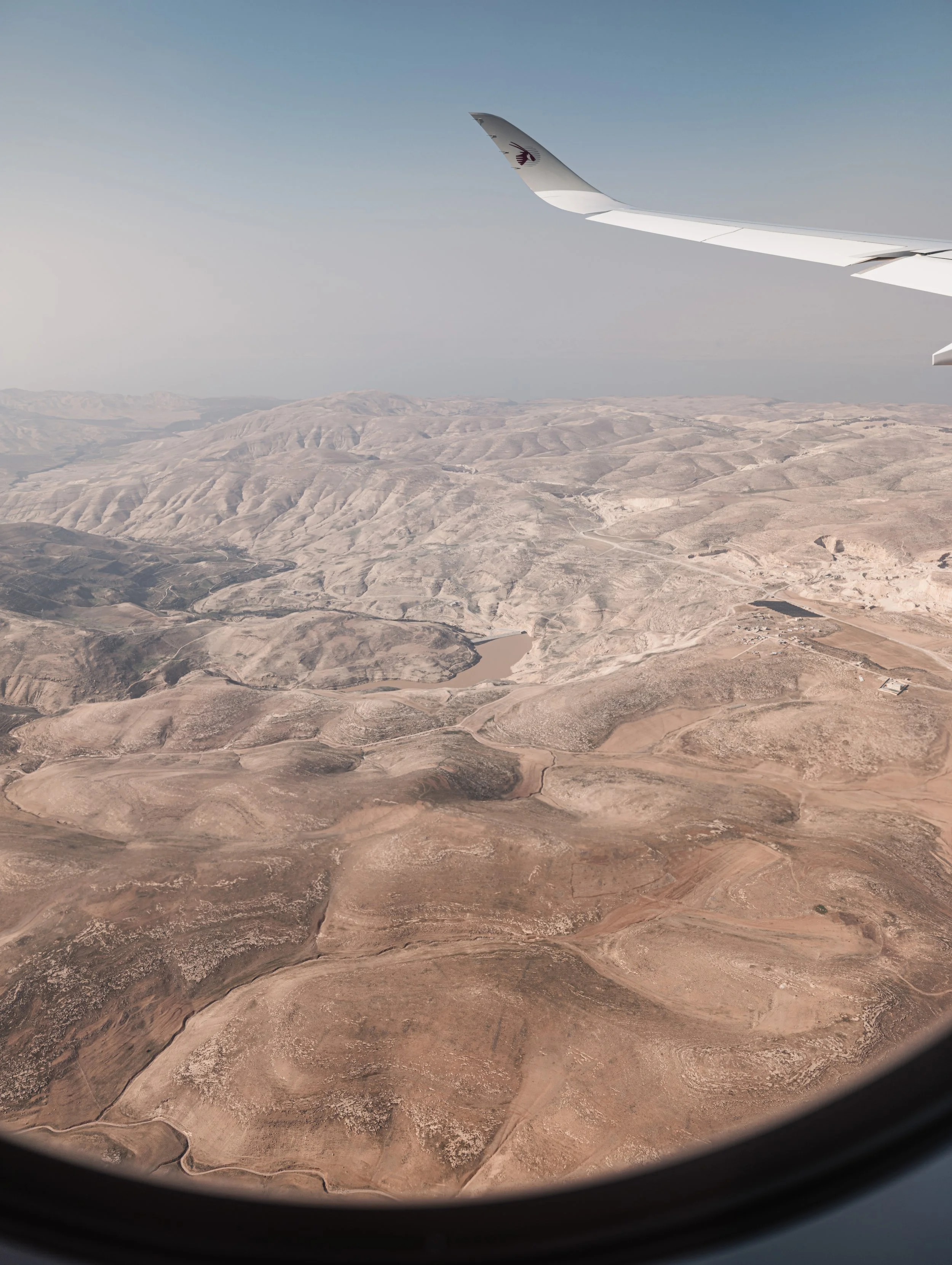View from an airplane window showing a desert landscape with hills and a body of water, with the plane's wing visible in the top right corner.