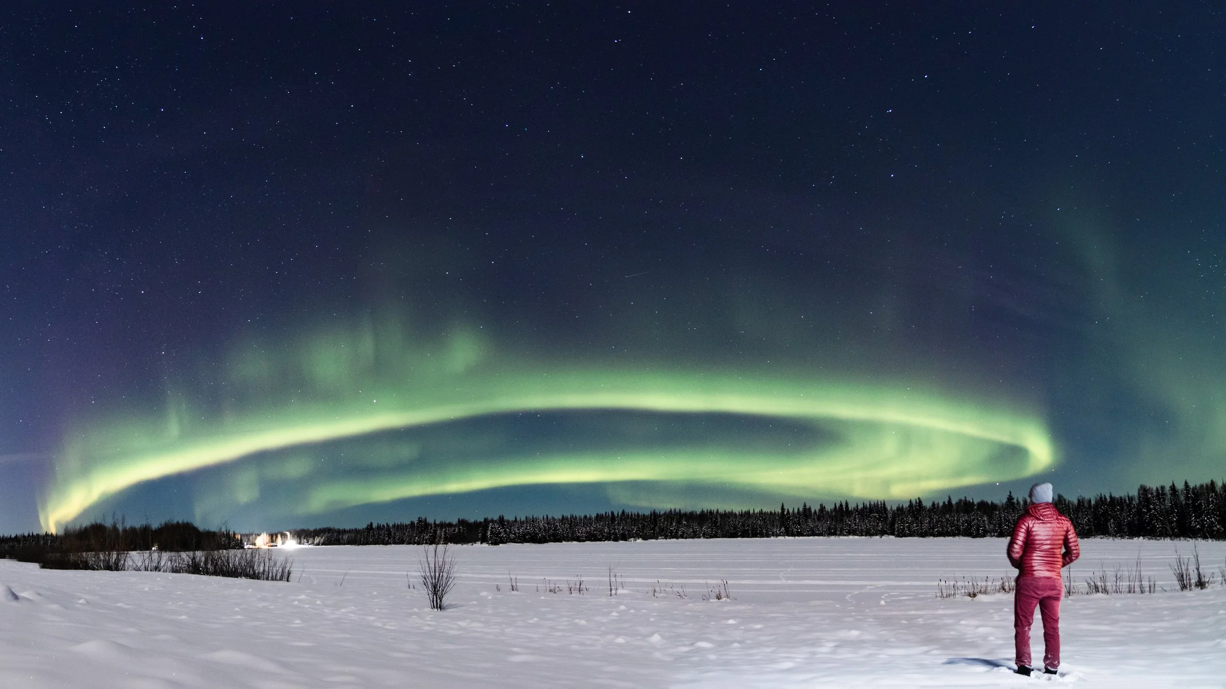 A person dressed in winter clothing standing on snow-covered ground, watching the northern lights in the night sky, which is filled with stars.