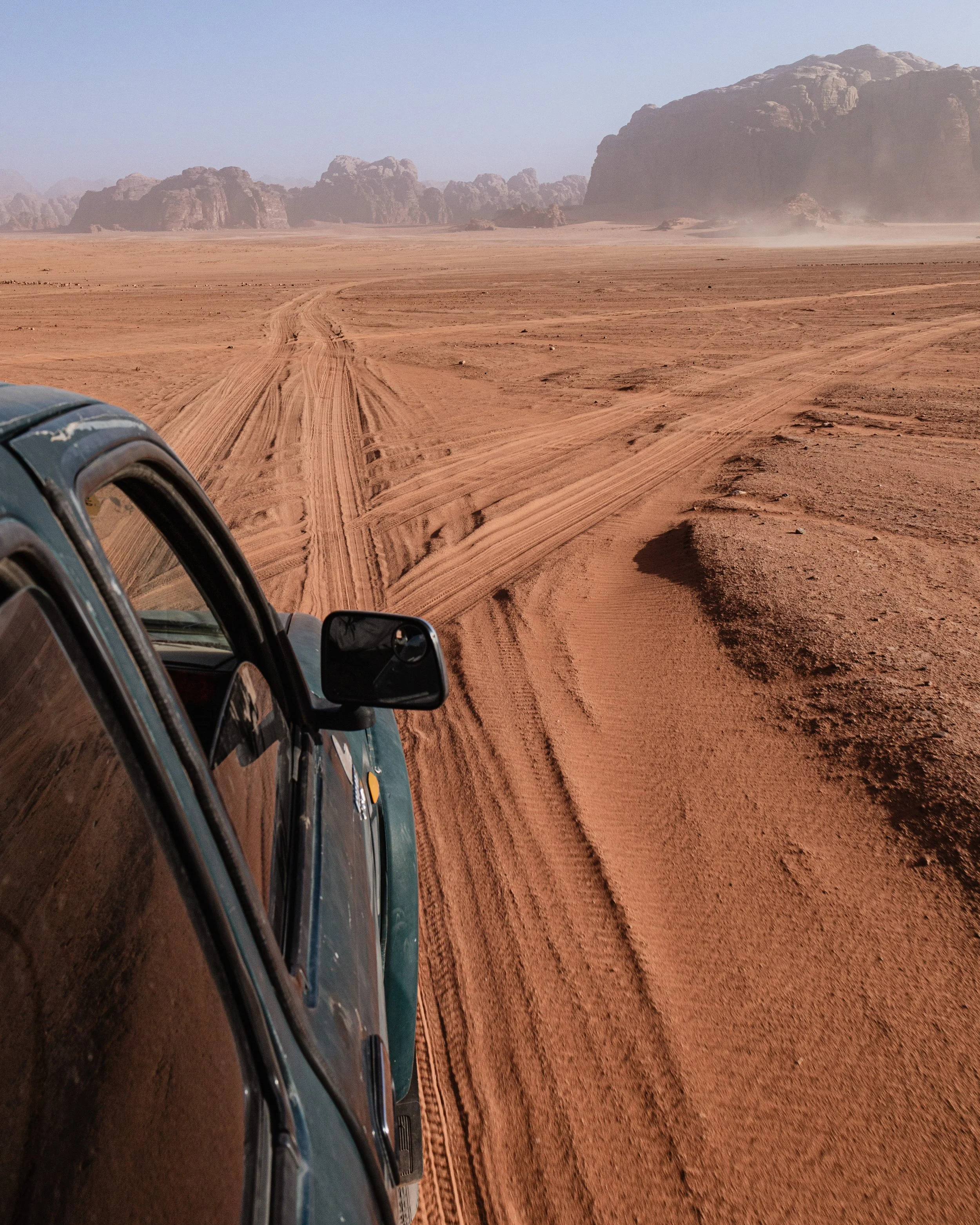 A vehicle driving through a desert landscape with reddish sand, rocky formations in the background, and tire tracks visible on the ground.
