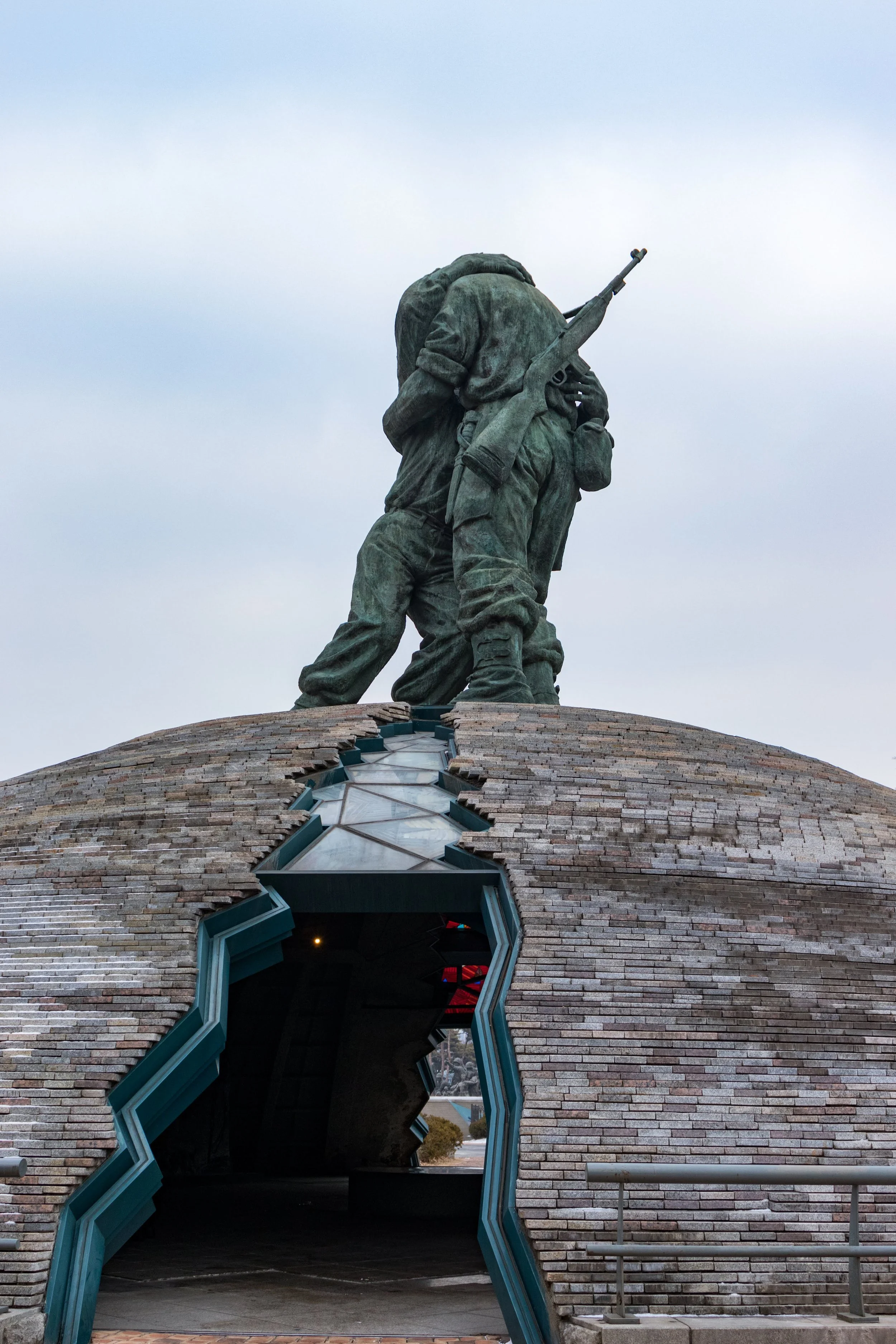 A monument with a sculpture of soldiers carrying a rifle, positioned on a brick structure with an irregular-shaped opening at its base.