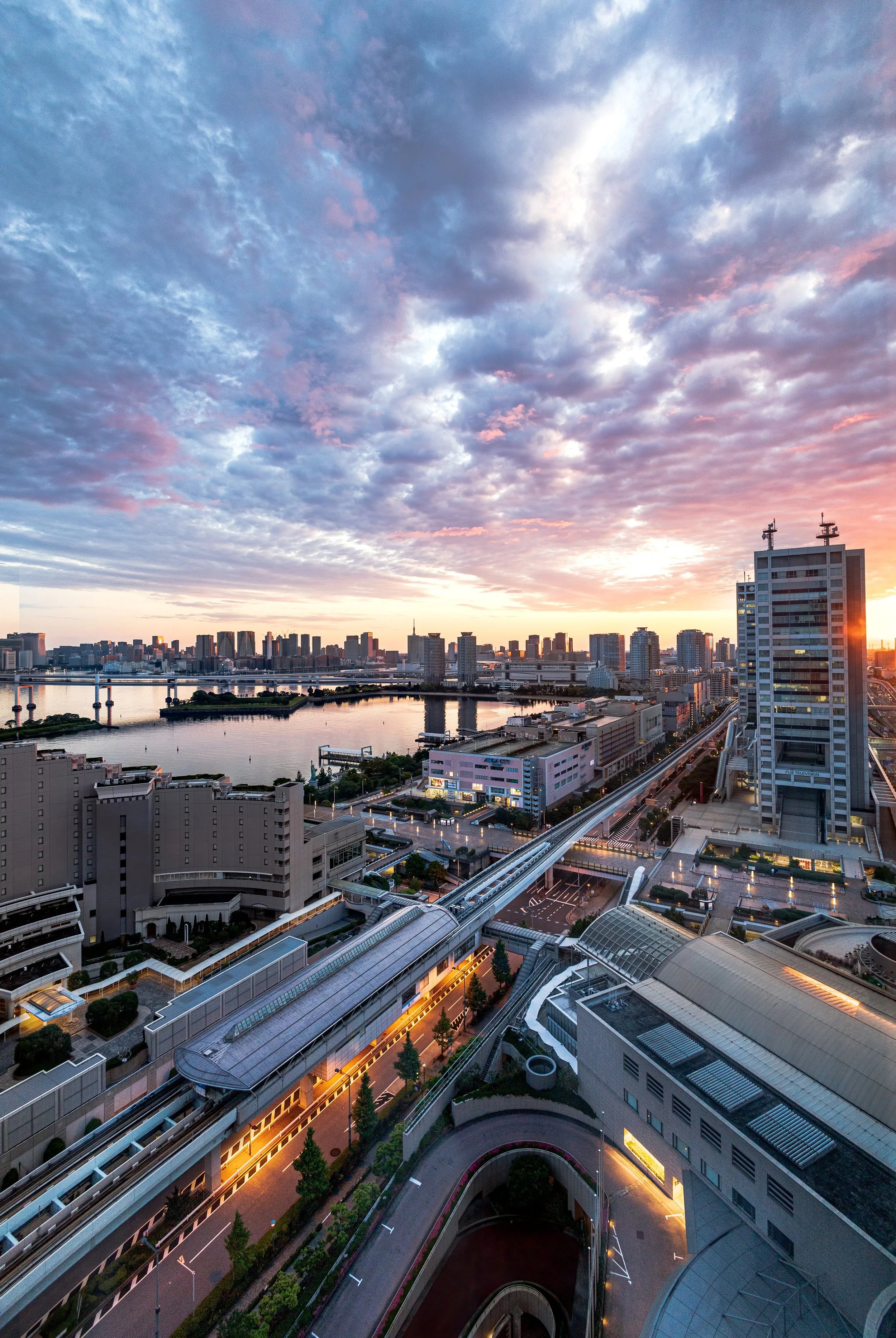 City skyline at sunset with tall buildings, a river, and a sky with orange and blue clouds.