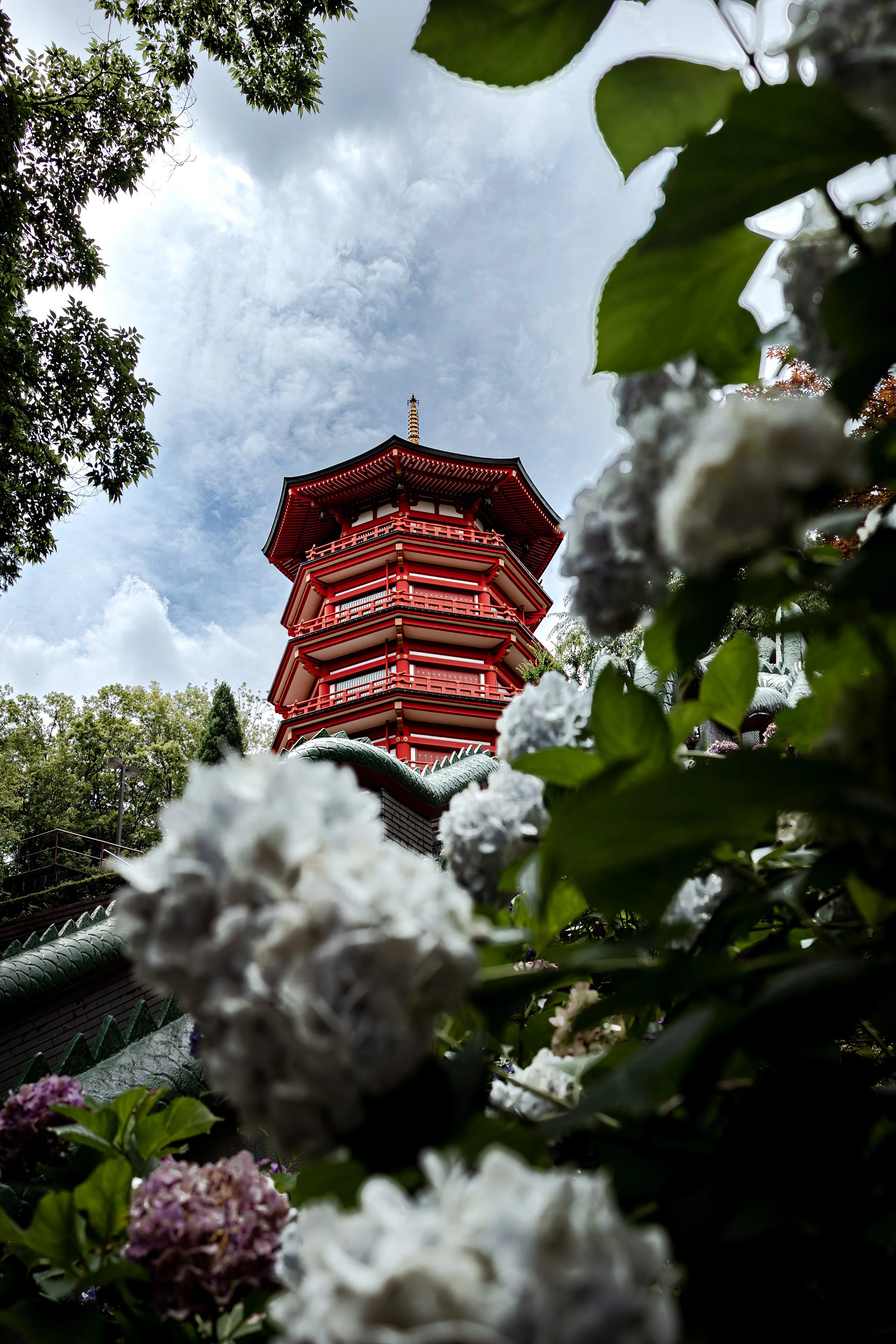 A traditional Japanese pagoda with red trim, viewed from below through white and purple flowers and green leaves, against a cloudy sky.