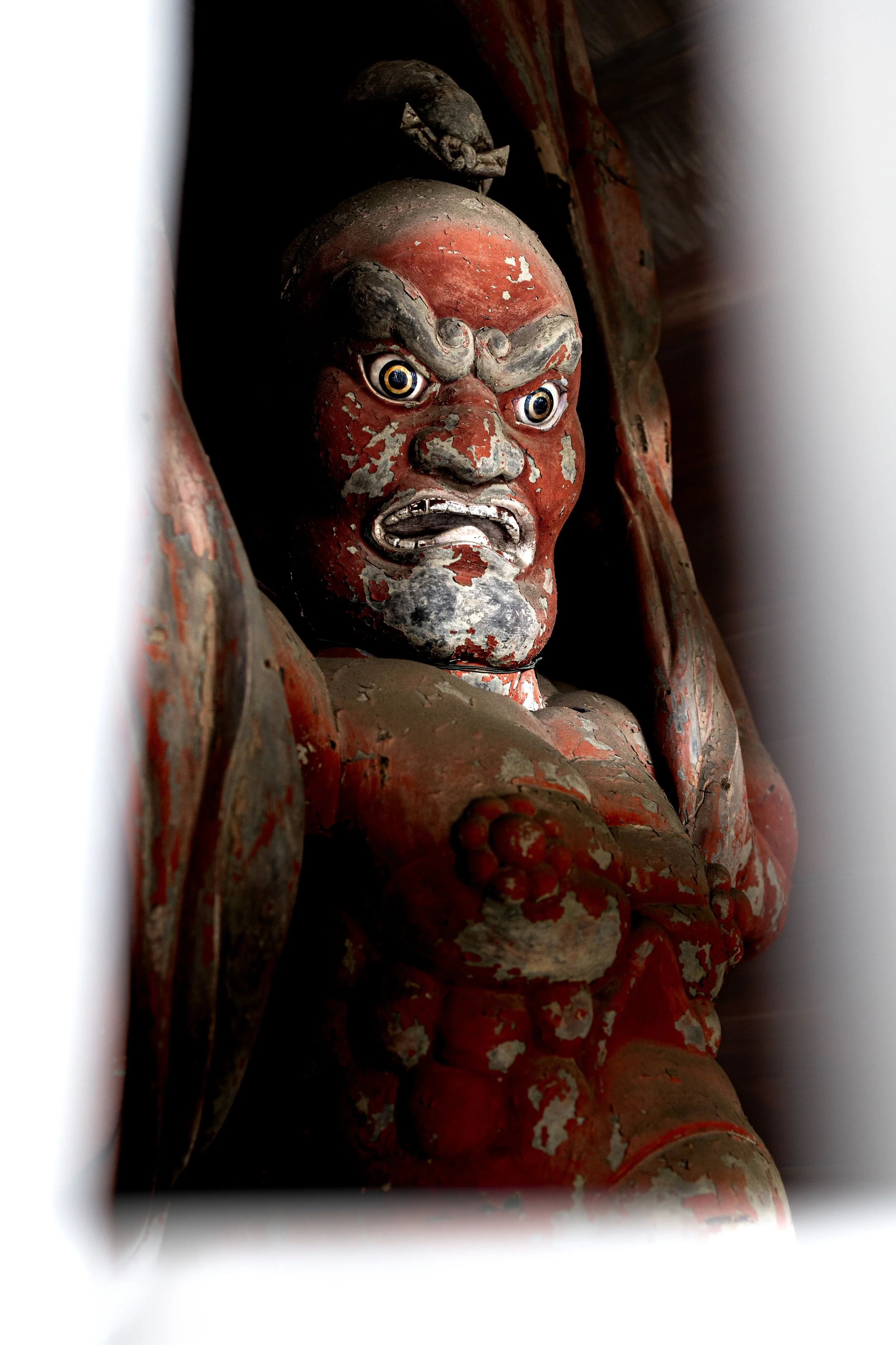 Close-up of an ancient, weathered wooden statue with a fierce expression, prominent eyes, and red and black paint that is peeling.