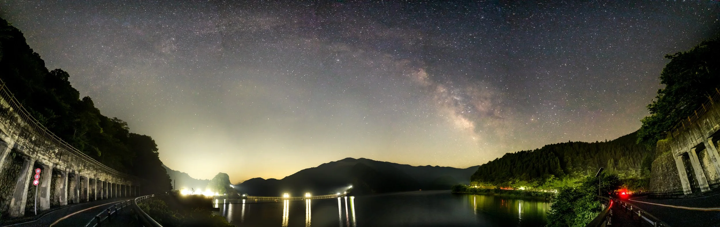 Night view of a starry sky with the Milky Way galaxy over a river surrounded by mountains, with a road and guardrails along the water, illuminated by distant lights and some vehicle lights.