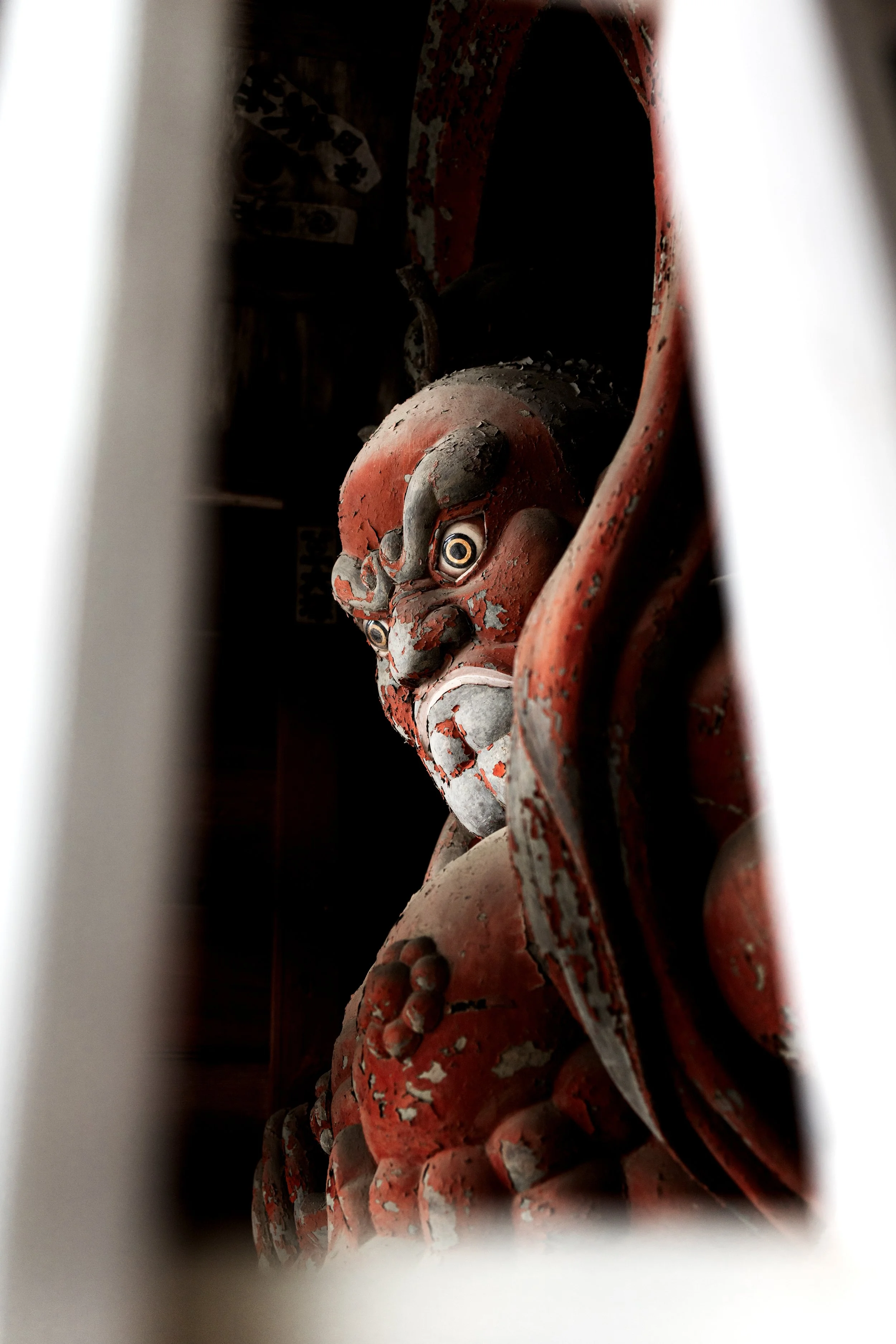 Close-up of a weathered, red and black painted, carved wooden face sculpture with intricate details and large, expressive eyes.