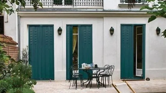 Patio avec une table et des chaises en fer forgé devant une maison blanche avec volets verts.