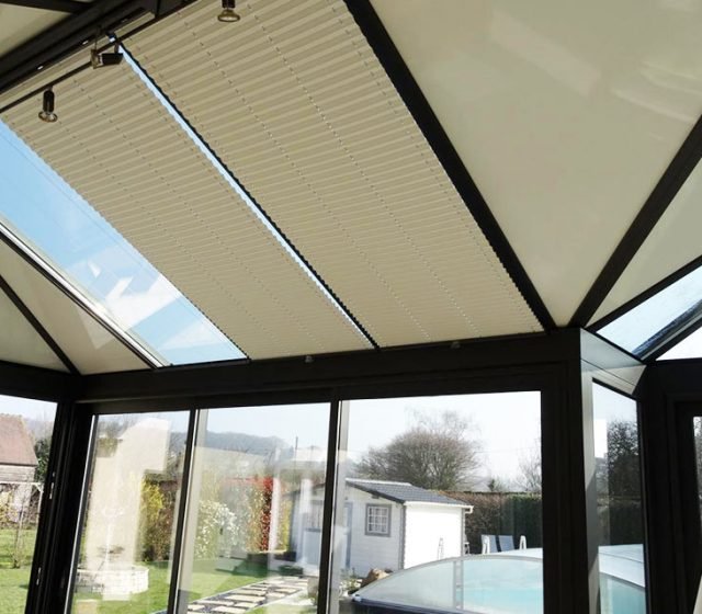 Une véranda avec des volets ou stores roulants partiellement ouverts au plafond, fenêtres en verre laissant voir un jardin extérieur.