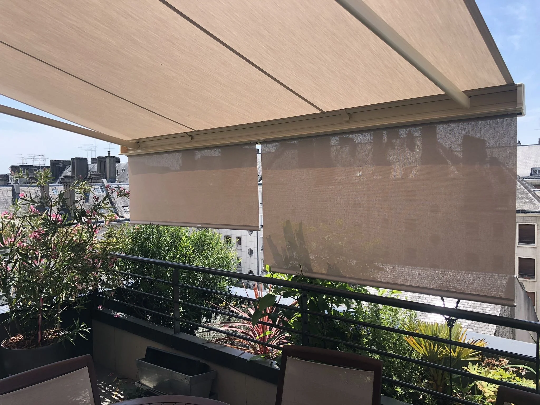 Balcon avec store parasol beige suspendu, vue sur des bâtiments avec baies vitrées et un ciel bleu, plantes en pot et meubles extérieurs dans le Rhône