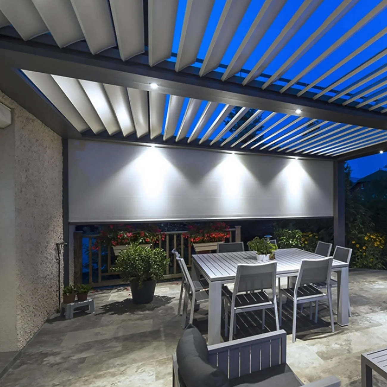 Terrasse extérieure couverte avec une table en bois gris, des chaises et des plantes décoratives, éclairée par des projecteurs modernes, en soirée.