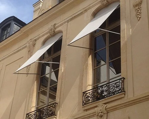 Deux fenêtres avec des volets en fer forgé et des auvents blancs sur un bâtiment en façade beige.