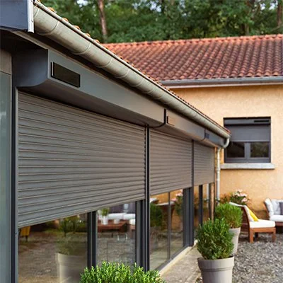 Une maison avec des volets roulants gris fermé devant une grande fenêtre, extérieur pavé avec des plantes en pot et un coin salon avec fauteuils en arrière-plan.