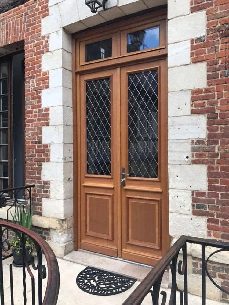 Porte en bois avec doubles battants, verre avec motif en croix, entourée de murs en briques et pierre, devant une entrée avec tapis en caoutchouc noir.