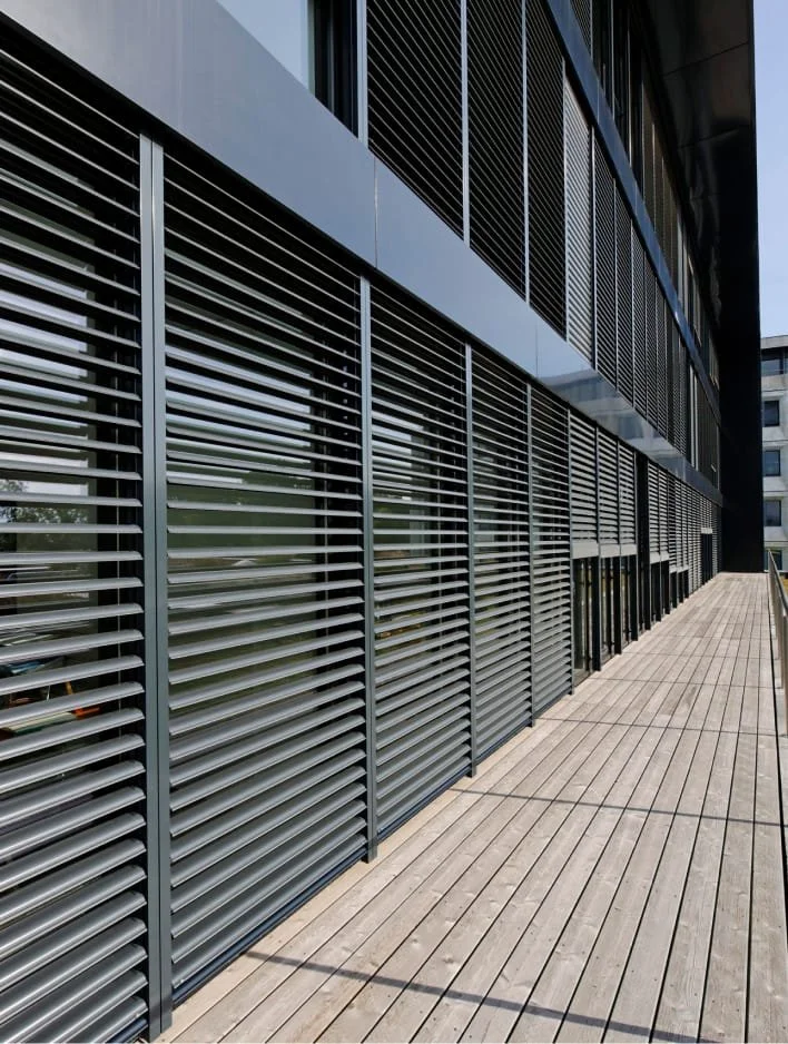 Façade d'un bâtiment moderne avec des volets en lattes métalliques et un balcon en bois.