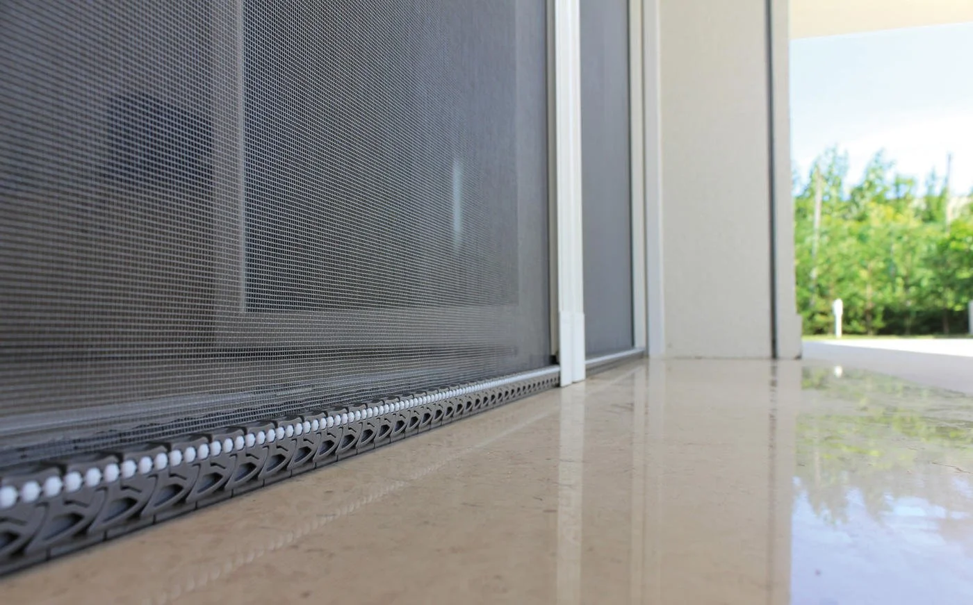 Vue au ras du sol d'une porte moustiquaire en métal avec un sol en marbre, à l'extérieur on voit des arbres verts et un ciel bleu.