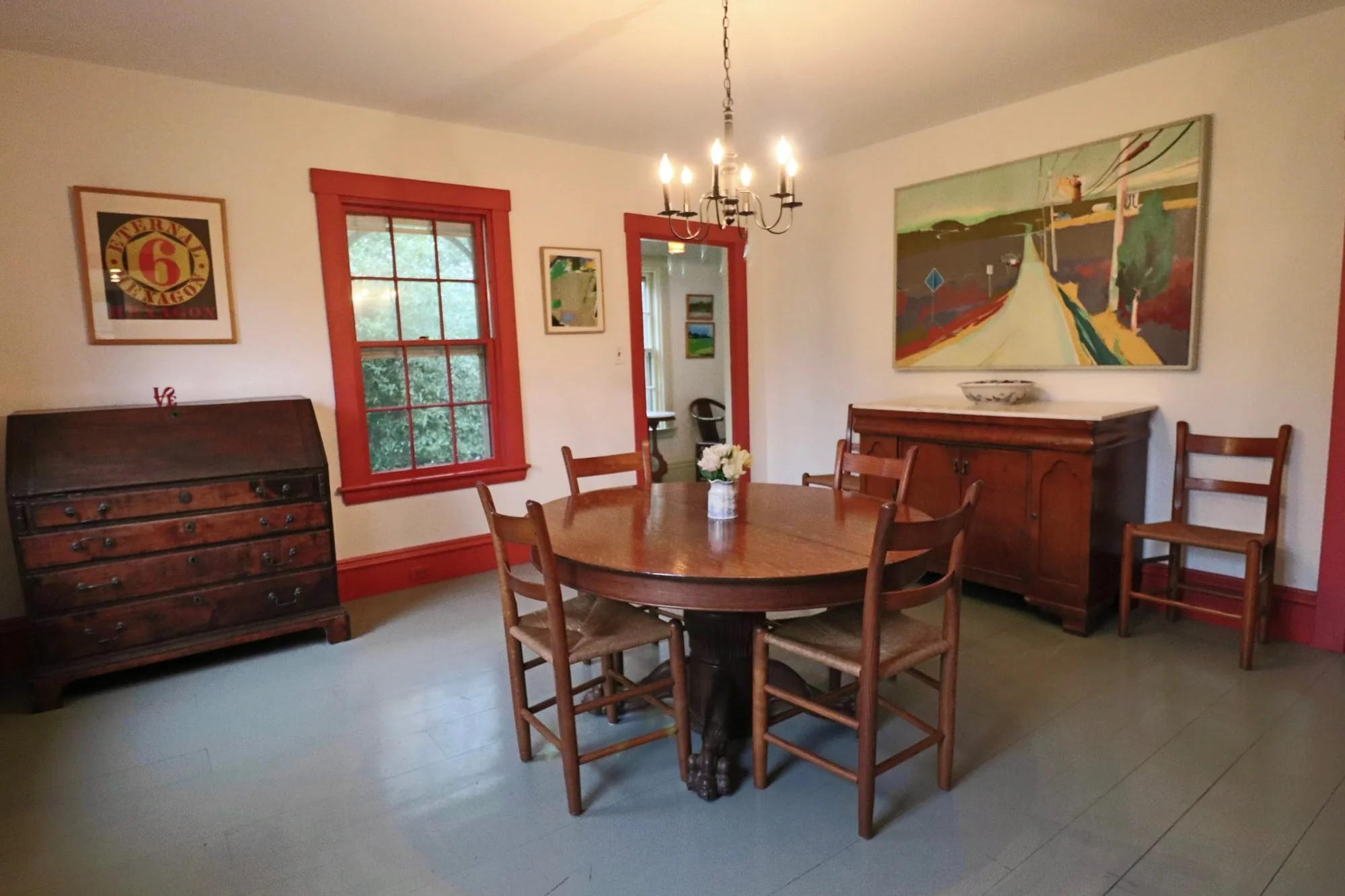Farmhouse dining room with a round antique table, red window trim and a chandelier in West Tisbury