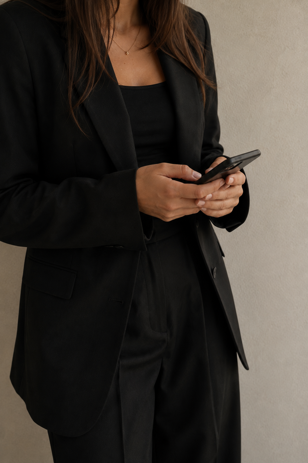 A woman dressed in a black blazer and black pants using a smartphone against a plain wall.