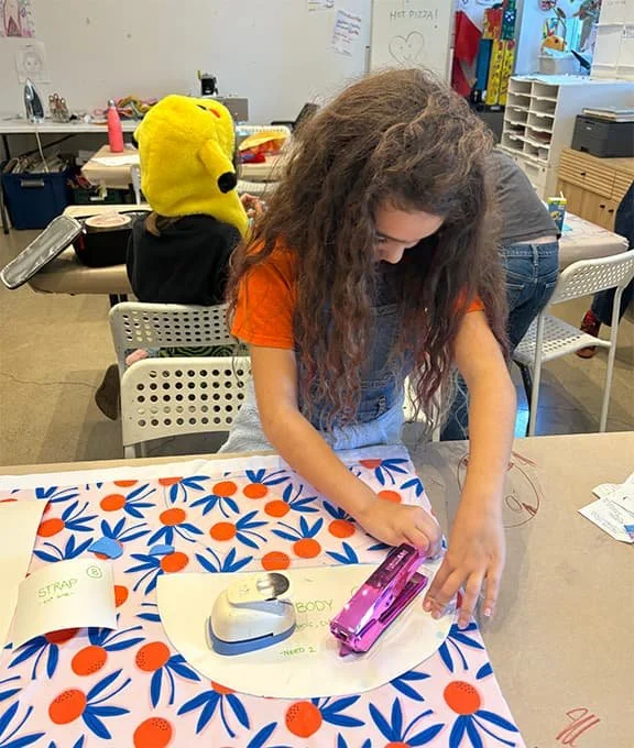 A girl with long curly hair and an orange shirt is stamping on a paper with a pink stamp, at a table covered with a colorful patterned paper. In the background, a child wearing a yellow Snoopy hat is sitting at a different table in a classroom.