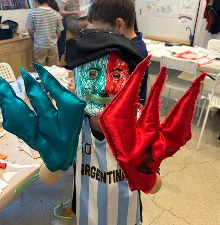 Child holding two Halloween-style gloves, one blue and one red, and a colorful mask with red, green, and black scribbles in a classroom setting.