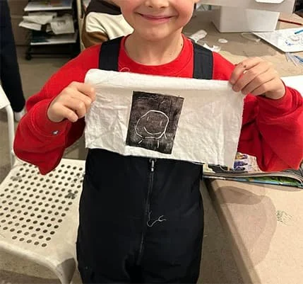 A young boy holding up a piece of paper with a chalk drawing of a circle and other shapes.