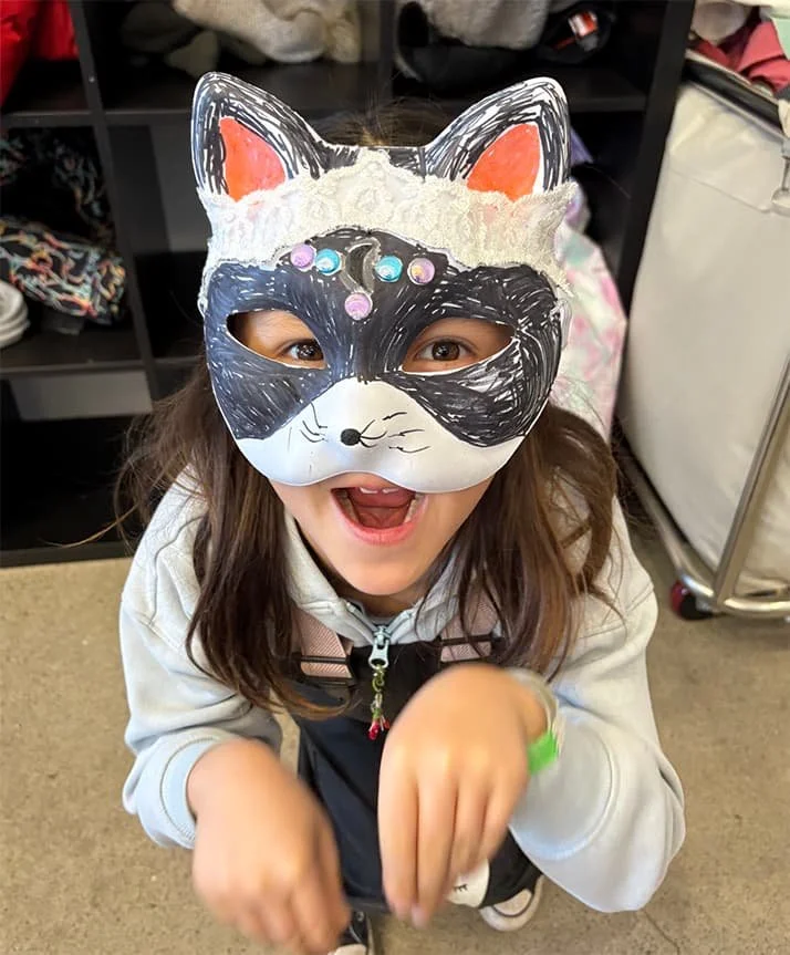 A smiling girl wearing a raccoon mask with multicolored beads and a white hoodie, making a playful paw gesture.