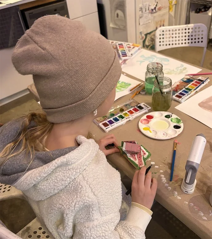 A young girl wearing a beige beanie and cozy hoodie sits at a cluttered table doing watercolors, with painting supplies and drawings around her.