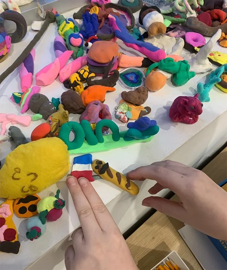 Colorful clay or playdough animals and objects on a table, including a yellow creature with a face, a French flag, and a giraffe, with hands in the frame.