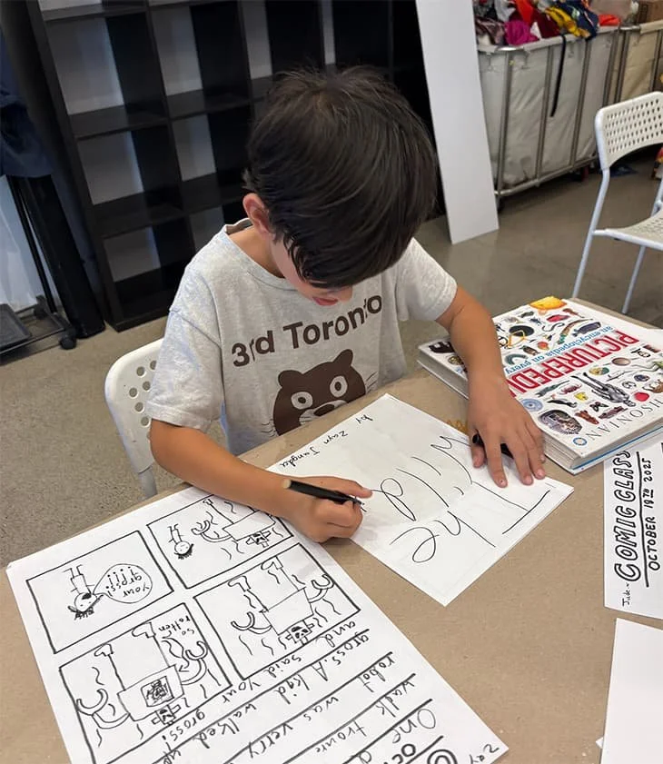 A young boy is sitting at a table, writing on a large sheet of paper with a marker. The paper has comic strip panels and handwriting on it. There are books and a chair nearby, and storage bins in the background.