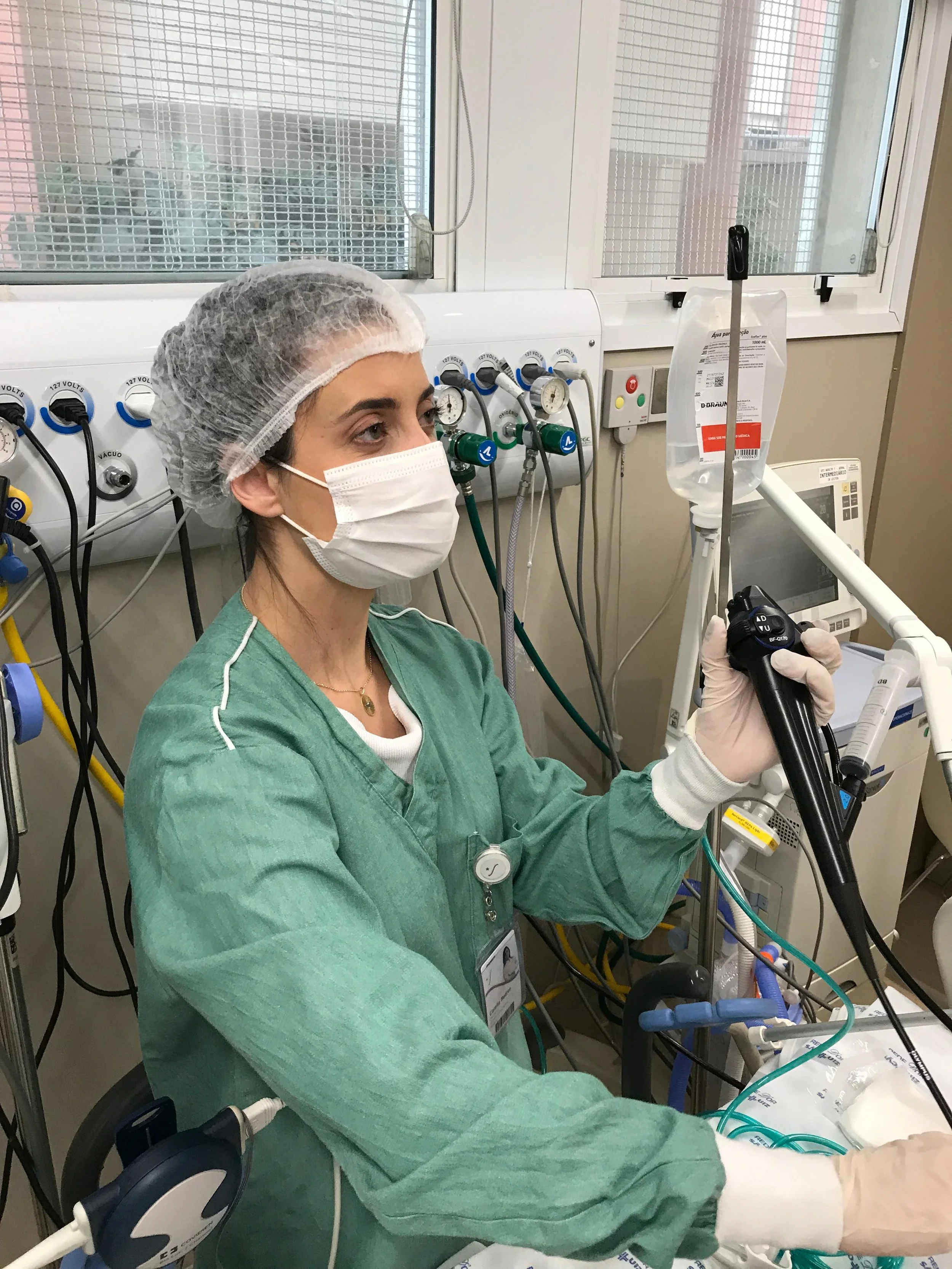 Médica em ambiente hospitalar realizando exame de endoscopia ou colonoscopia. Ela veste scrubs verdes, máscara cirúrgica, touca e luvas, manuseando um endoscópio. Ao fundo, painel com gases medicinais e monitor hospitalar.