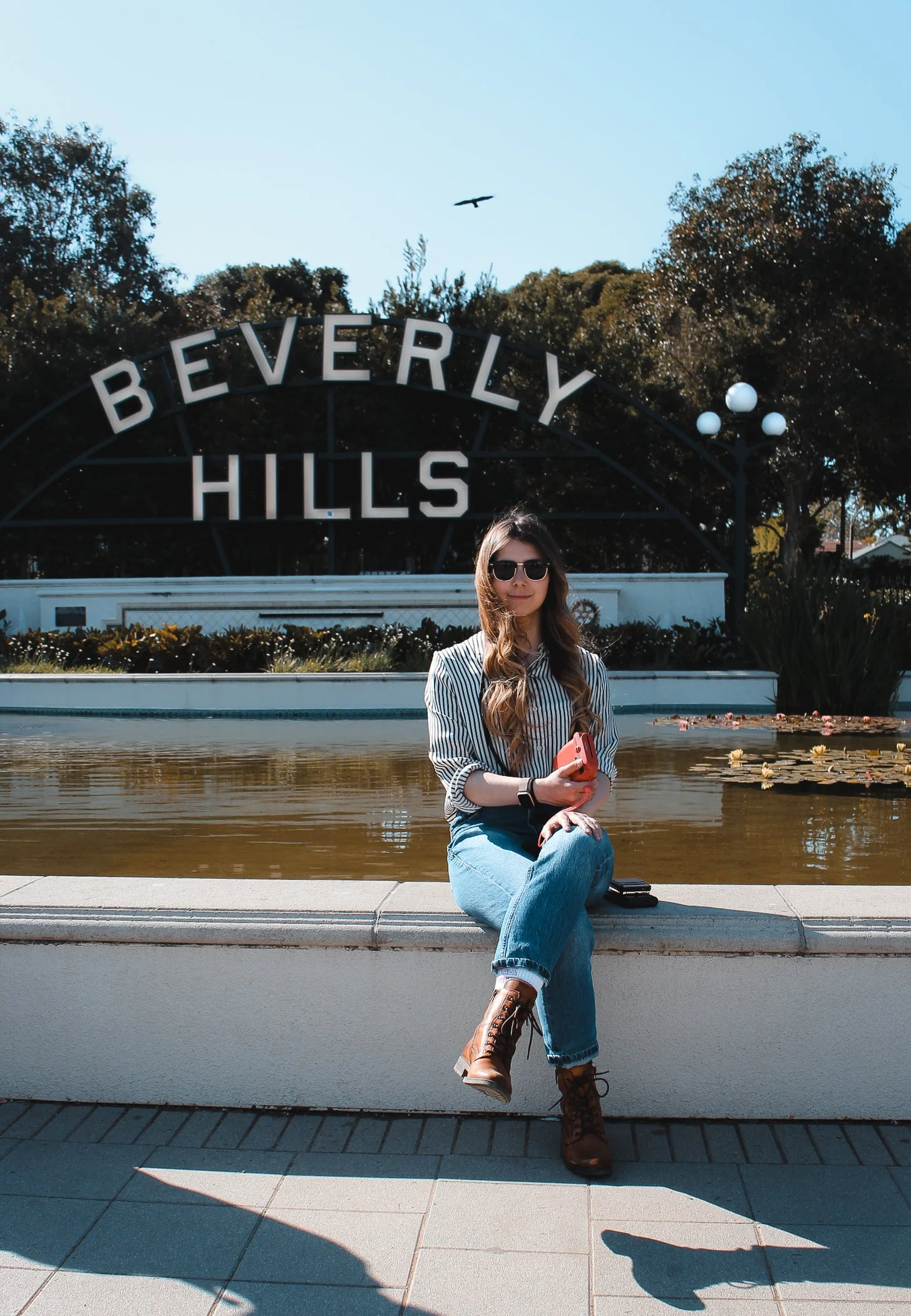 A young woman sitting on a ledge in front of a pond and a sign that reads 'Beverly Hills.' She is wearing sunglasses, a striped shirt, jeans, and brown lace-up boots, and is holding a pink phone.