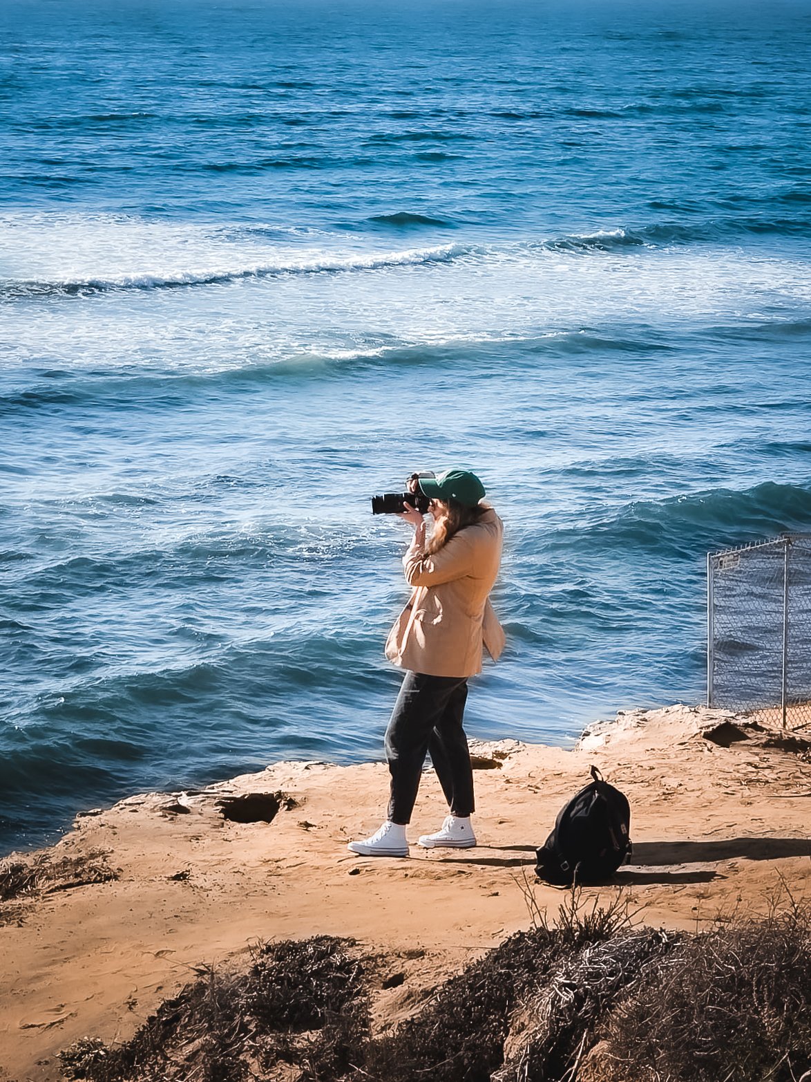 A person taking a photo with a camera on a rocky shoreline next to the ocean, wearing a beige jacket, black pants, white sneakers, and a green hat, with a black backpack on the ground nearby.