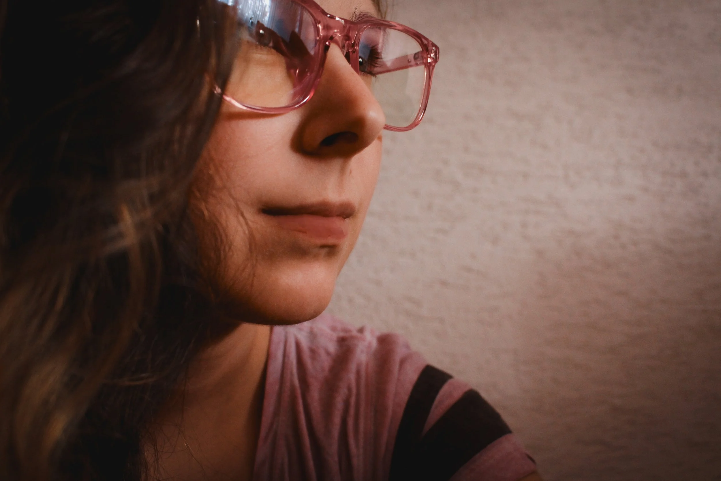A woman with long brown hair and pink glasses looking downward against a neutral background.