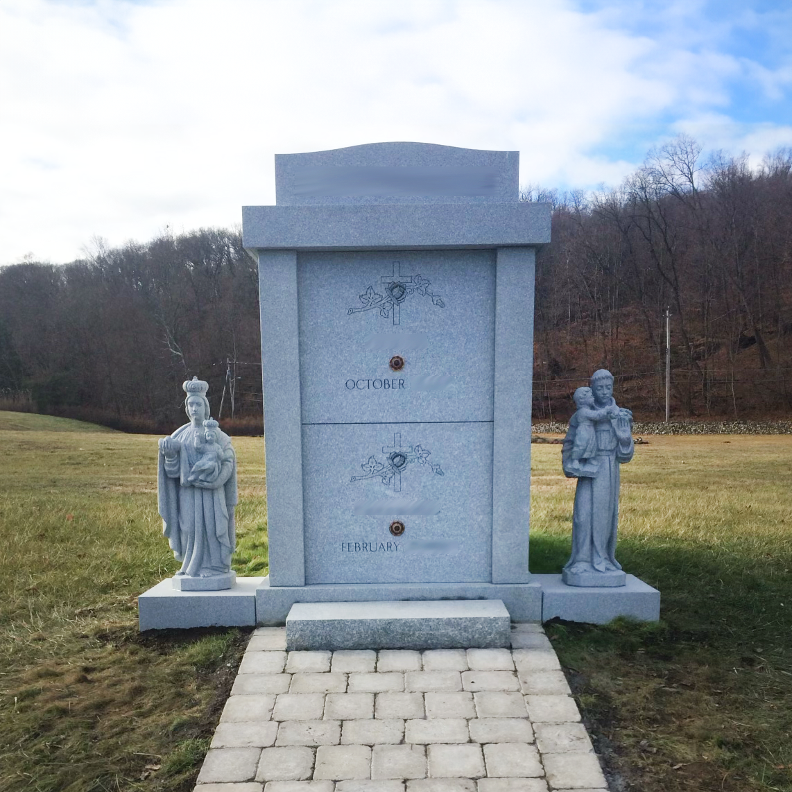 A gray granite mausoleum memorial with engraved religious symbols, flanked by two statues of saints.