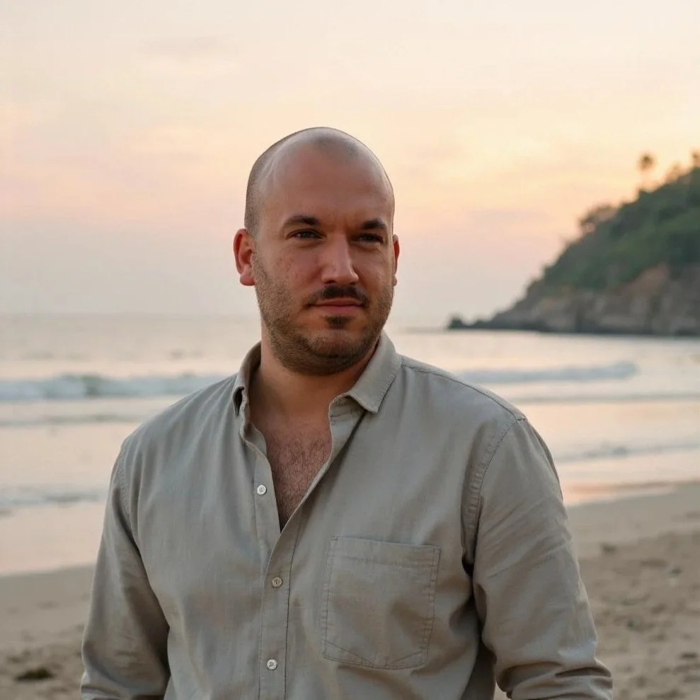 Mann mit Glatze und Bart am Strand bei Sonnenuntergang, Blick aufs Meer und eine Küste im Hintergrund.