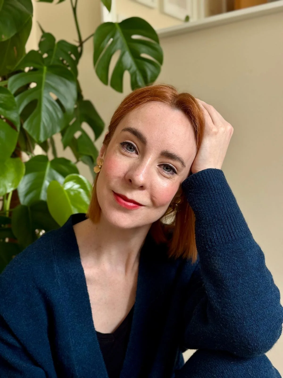 A woman with red hair, blue eyes, and fair skin, wearing a navy blue sweater and red lipstick, posed with her head resting on her hand, sitting in front of a large green plant with split leaves.