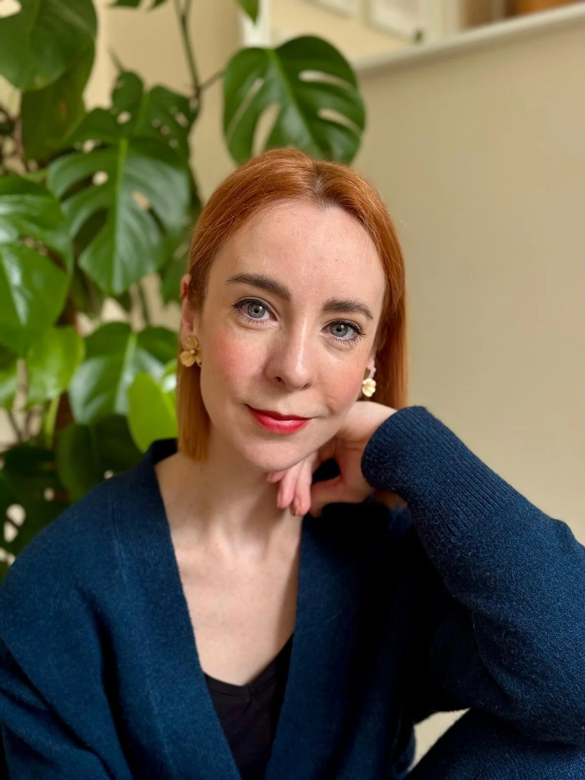 A woman with fair skin, red hair, blue eyes, wearing gold flower earrings and a dark blue cardigan, sitting indoors near a large green plant with broad leaves, resting her chin on her hand, looking at the camera.