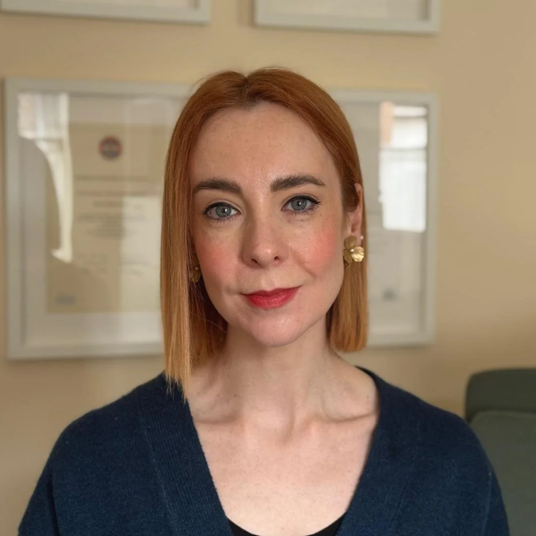 Portrait of a woman with red hair and blue eyes, wearing gold earrings and a dark blue sweater, in an indoor setting with framed documents in the background.