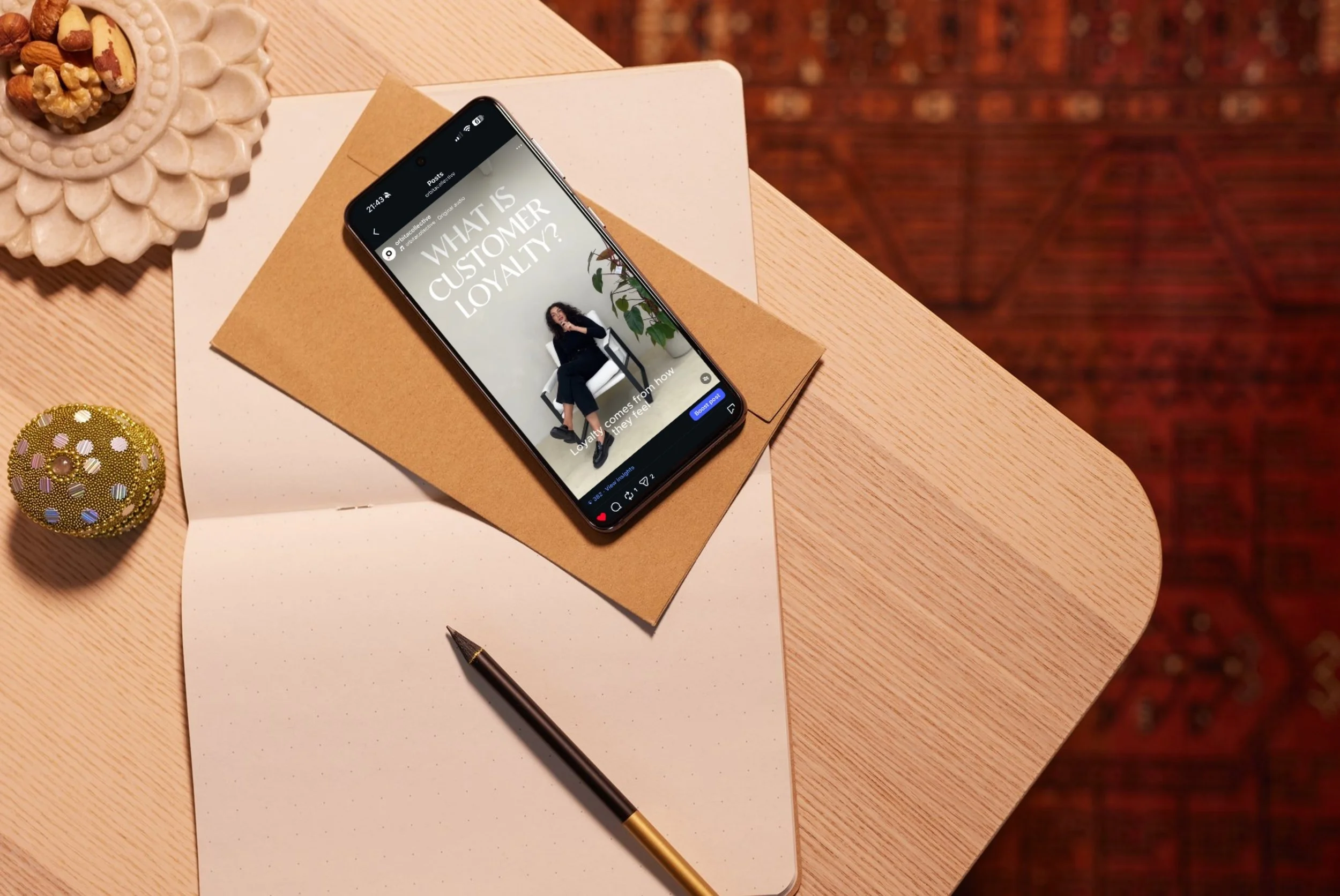 A smartphone displaying a social media post with the question 'What is customer loyalty?' placed on a brown and white notepad on a wooden table. Nearby are a black and gold pen, a white ceramic dish with snacks, and a gold decorative ball. The backgr