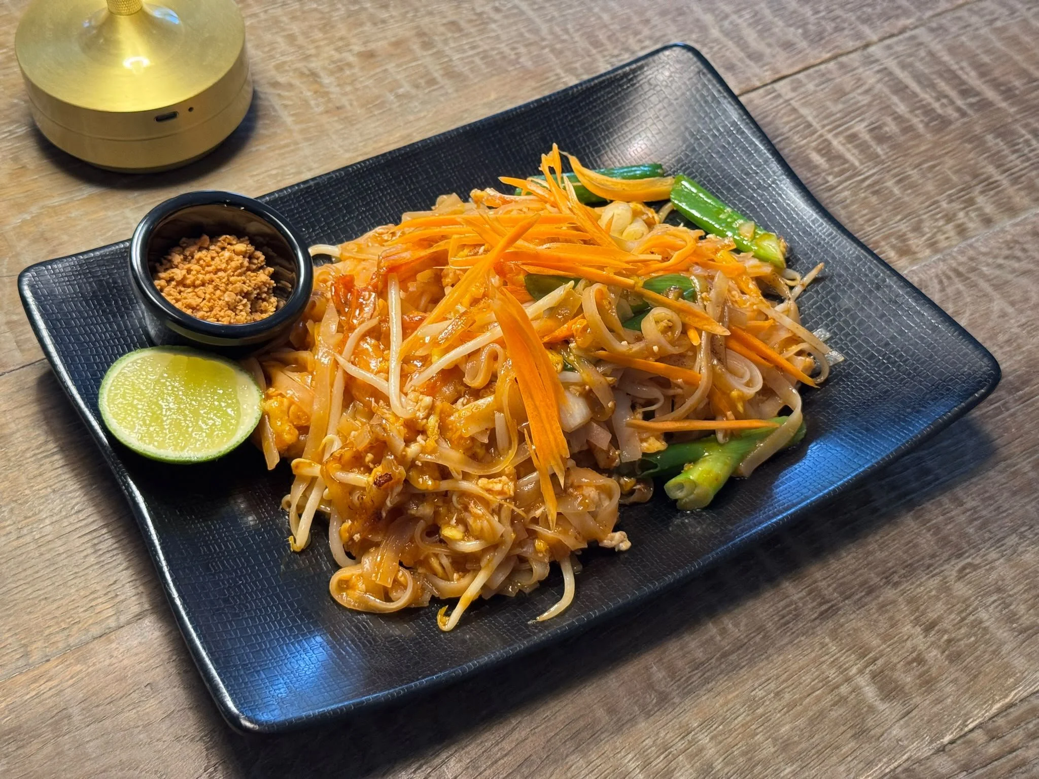 A black square plate with cooked Asian-style stir-fried noodles, sliced vegetables including green beans, carrots, and bean sprouts, a lime wedge, and a small black bowl of crushed peanuts. A yellow lamp is partially visible on a wooden table.