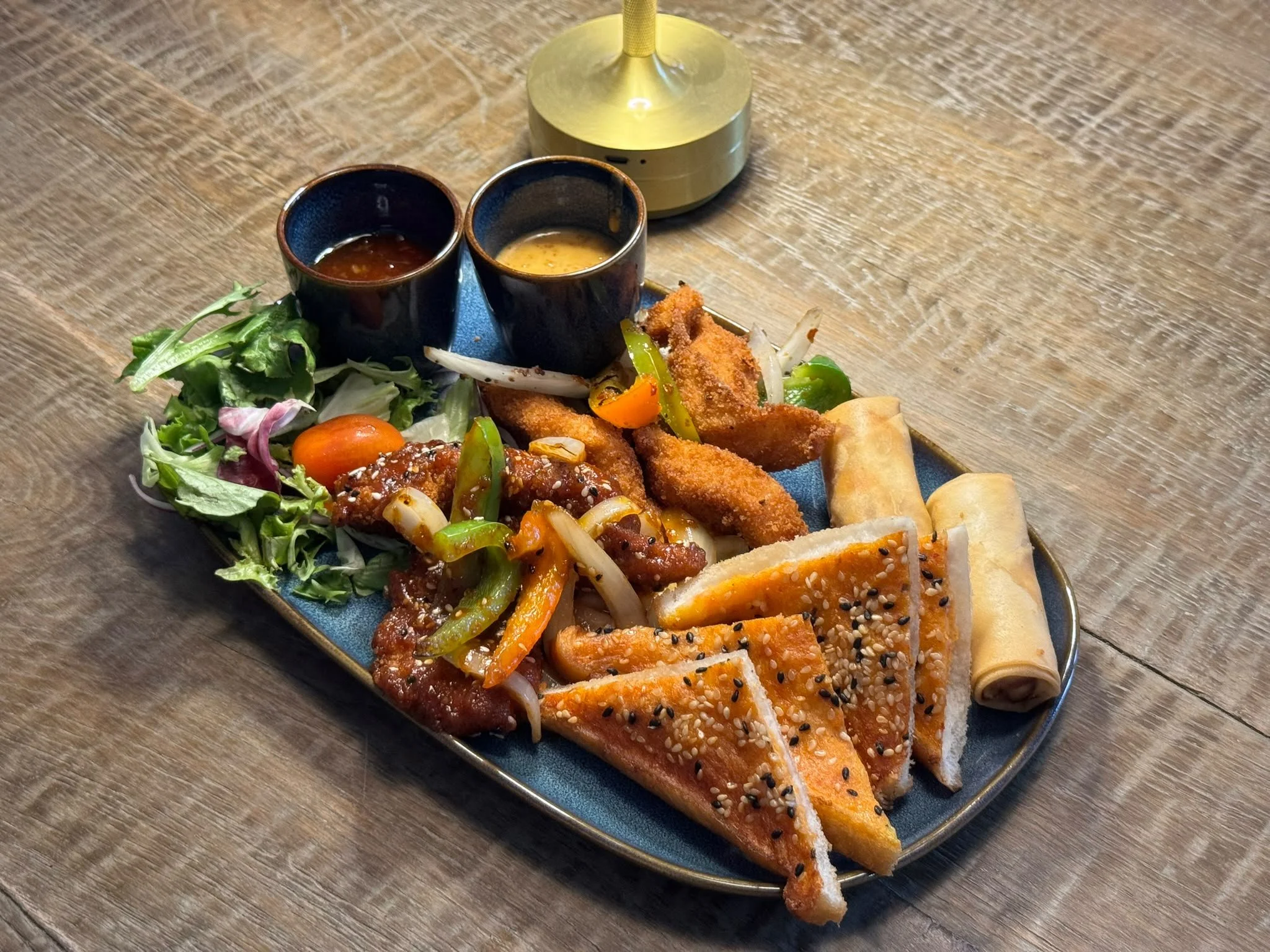 A long oval plate with an assortment of Asian appetizers on a wooden table. Includes salad greens, fried breaded fish fillets with orange sauce, spring rolls, and sesame seed-crusted toast slices. Two small black soy sauce dishes are also on the plate, one with dark sauce and the other with a yellow egg mixture. A gold-colored lamp is visible above.