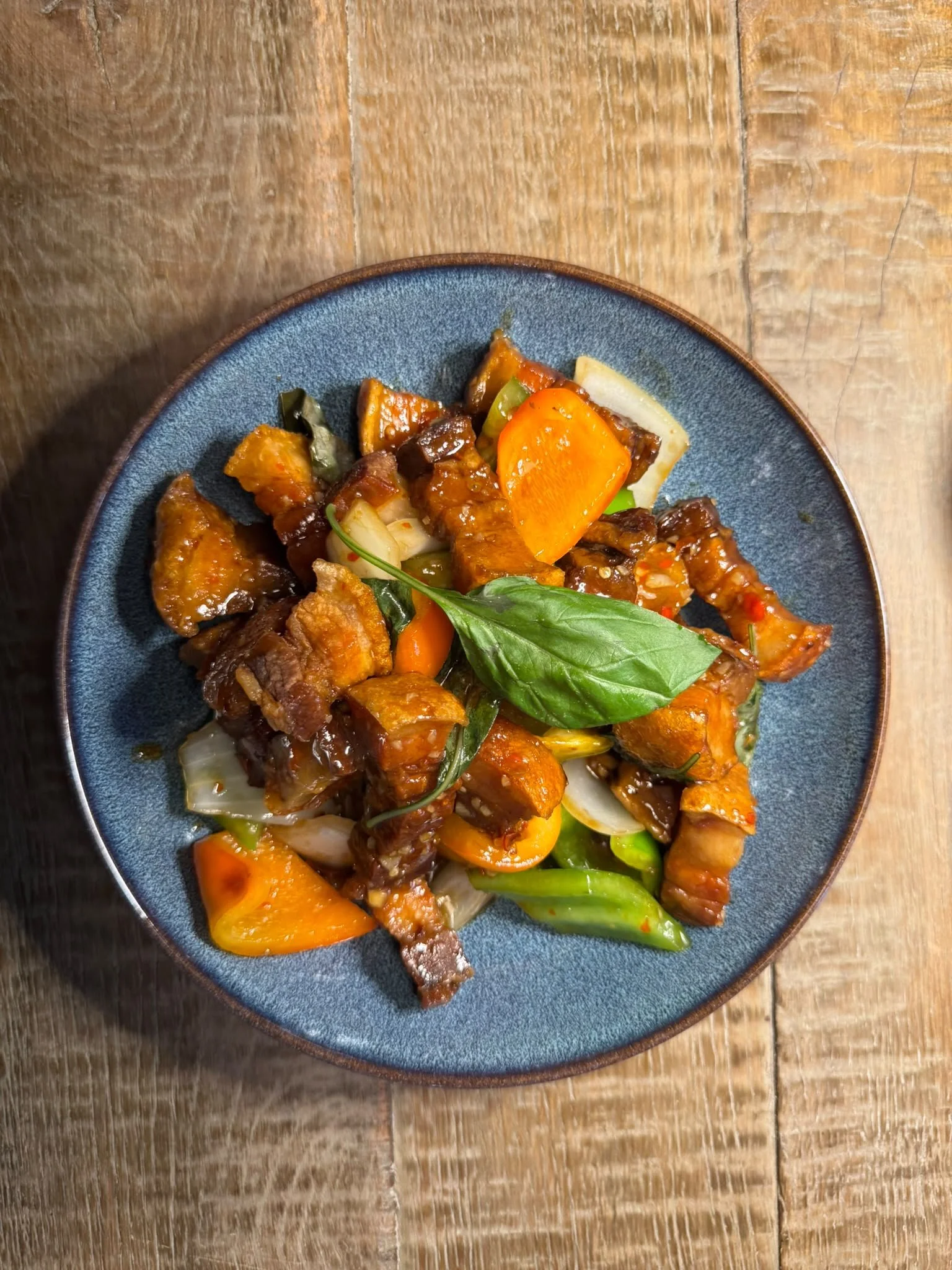 A plate of stir-fried vegetables and meat in a dark sauce on a blue ceramic plate, on a wooden table.