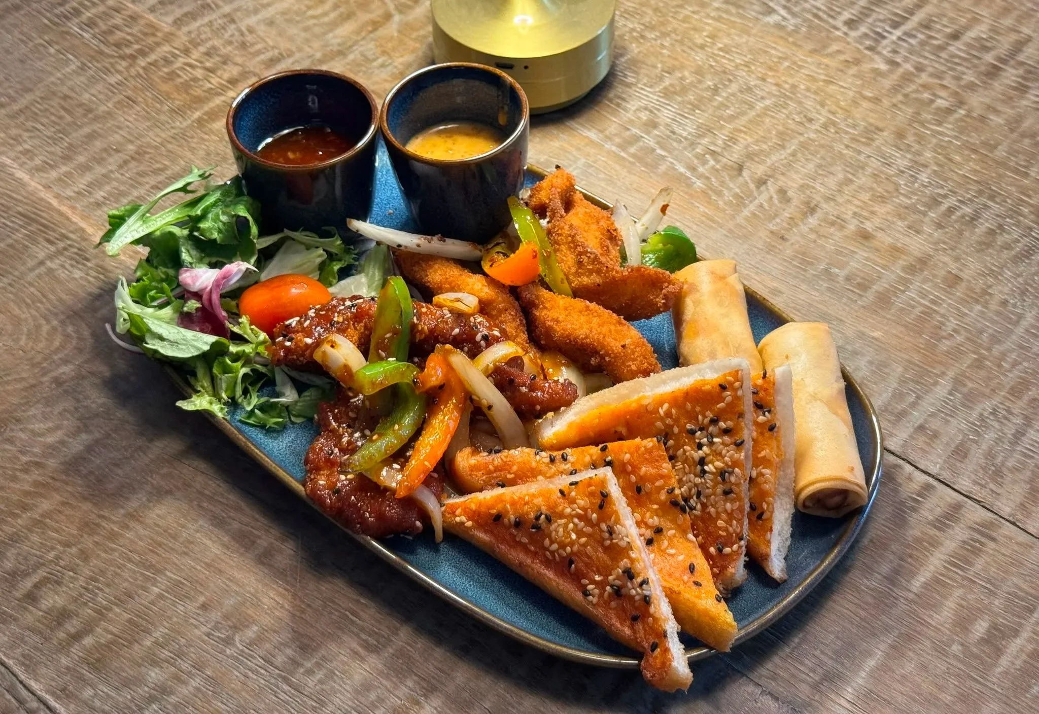 A rectangular plate with assorted Asian appetizers and dipping sauces on a wooden table.